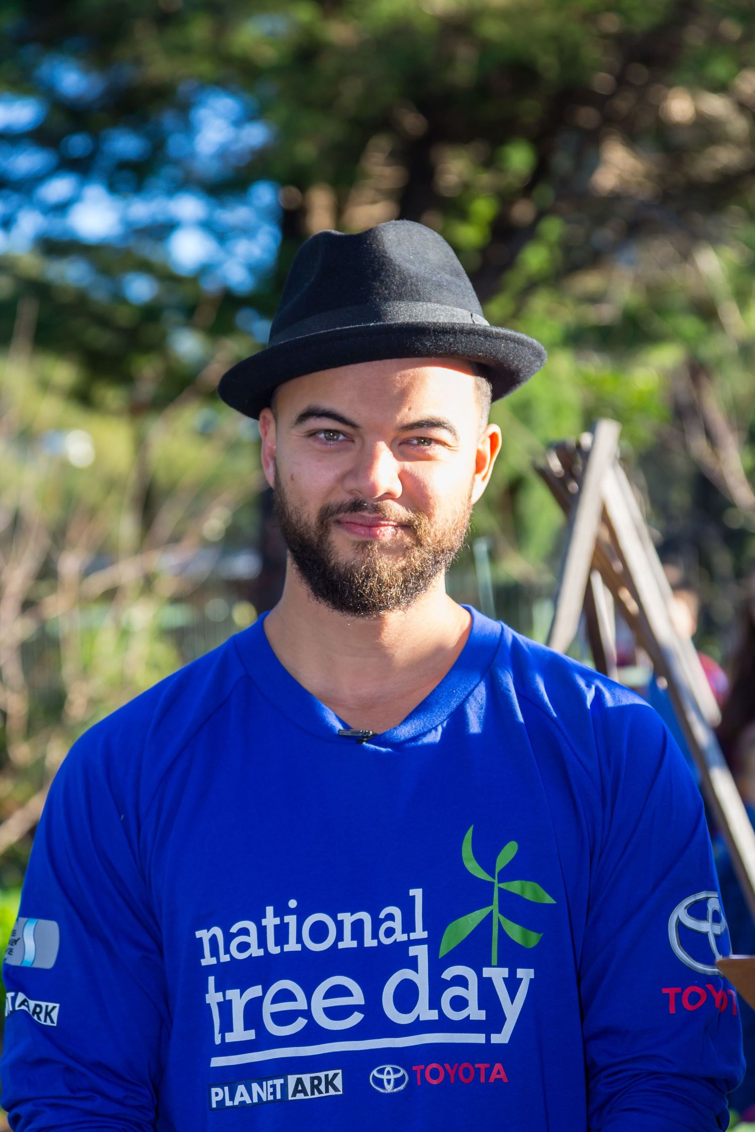 Bearded man in a black fedora and blue National Tree Day shirt smiles outdoors among trees at a community planting event.