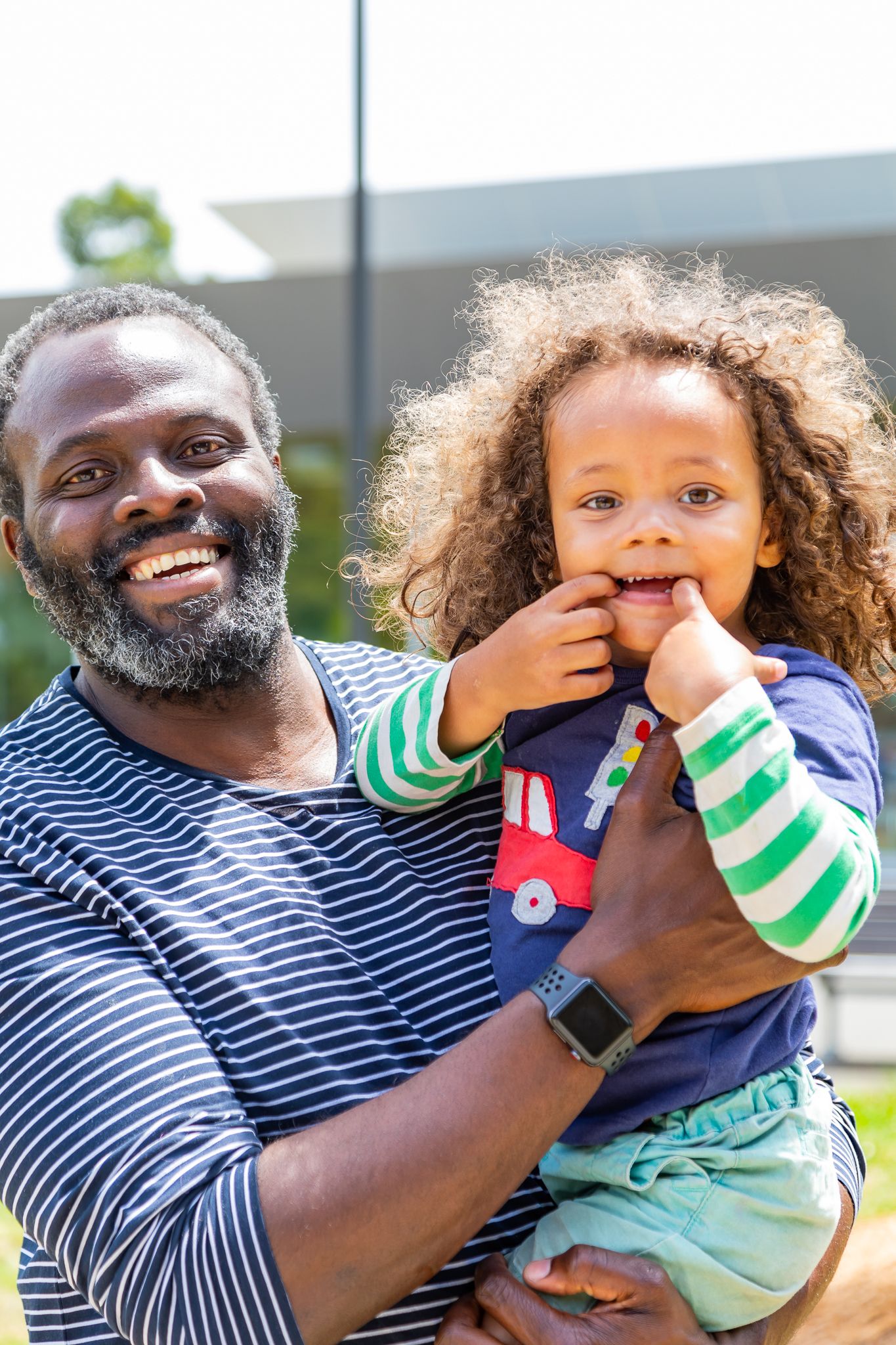 A bearded man in a striped shirt smiles while holding a curly-haired toddler who has fingers in his mouth.