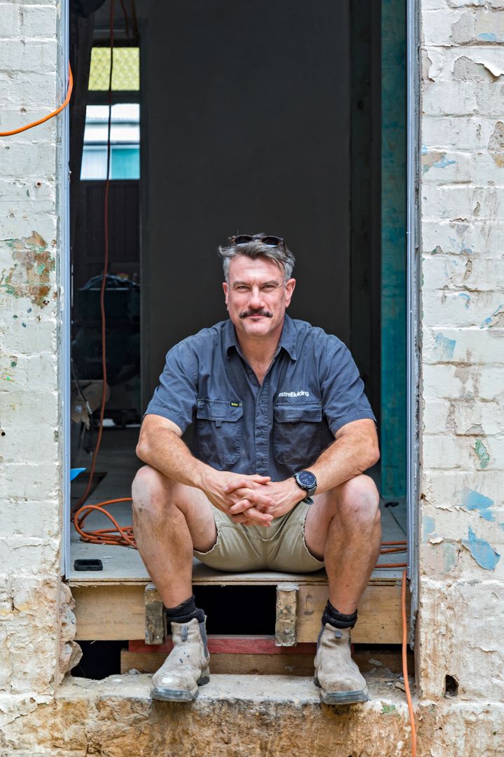 Smiling man with a moustache and sunglasses on his head sits in a doorway of a brick building under renovation wearing work boots.