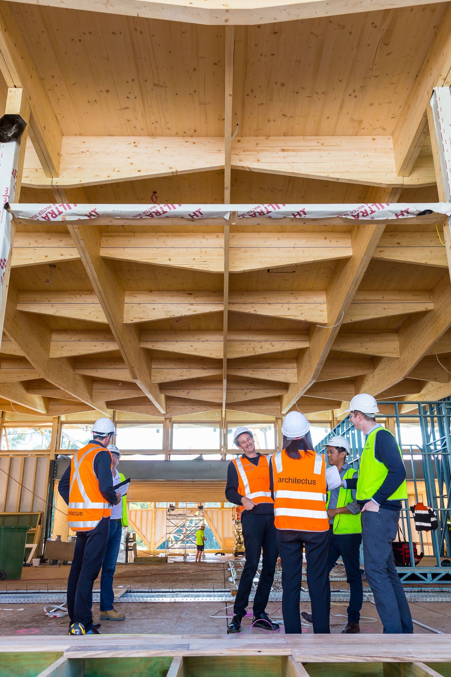 Construction workers in hi-vis vests and hard hats look up at exposed timber beams and a wooden ceiling in a building under construction.