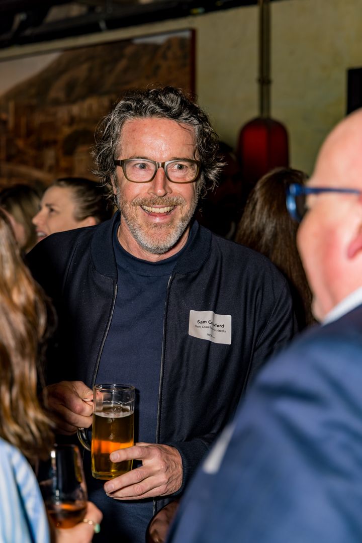 A smiling bearded man in glasses holds a glass of beer while chatting with guests at a social event.