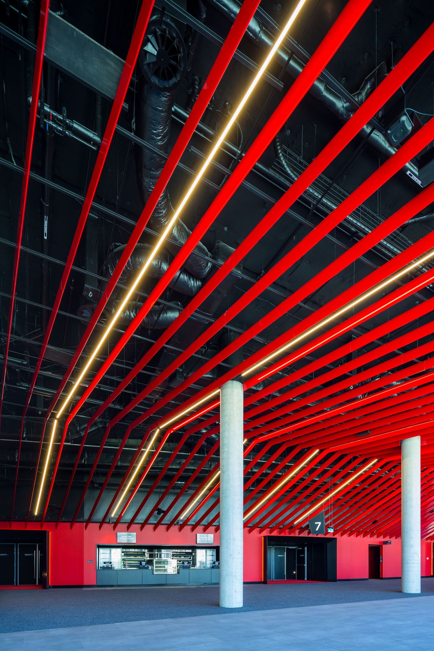 Covered walkway with bold red ceiling beams, glowing linear lights and white concrete columns leading to a food and beverage counter.