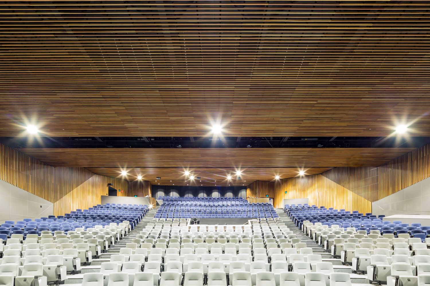 Large auditorium with blue and white seating, warm timber slat ceiling and wood-panelled walls under bright lights.