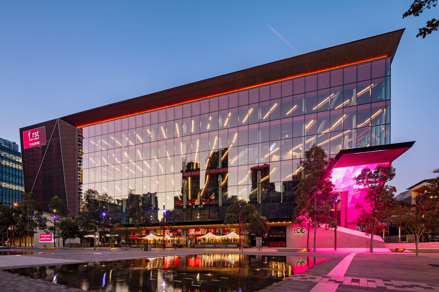 Modern glass convention centre with an illuminated orange roofline glowing at dusk beside a reflective pool and pink-lit trees.