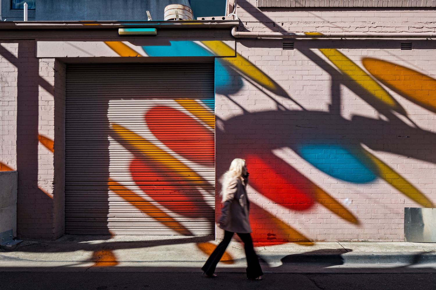 Pedestrian walks past a brick wall and roller door painted with bold red, blue and yellow abstract shapes.