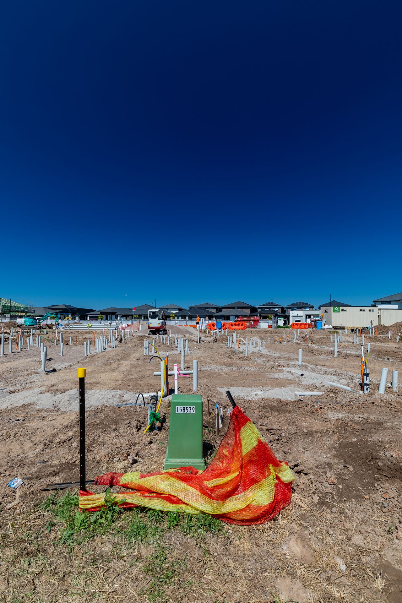 Green utility pedestal and orange safety netting on bare dirt at a housing development site with pipe stubs under a clear blue sky.