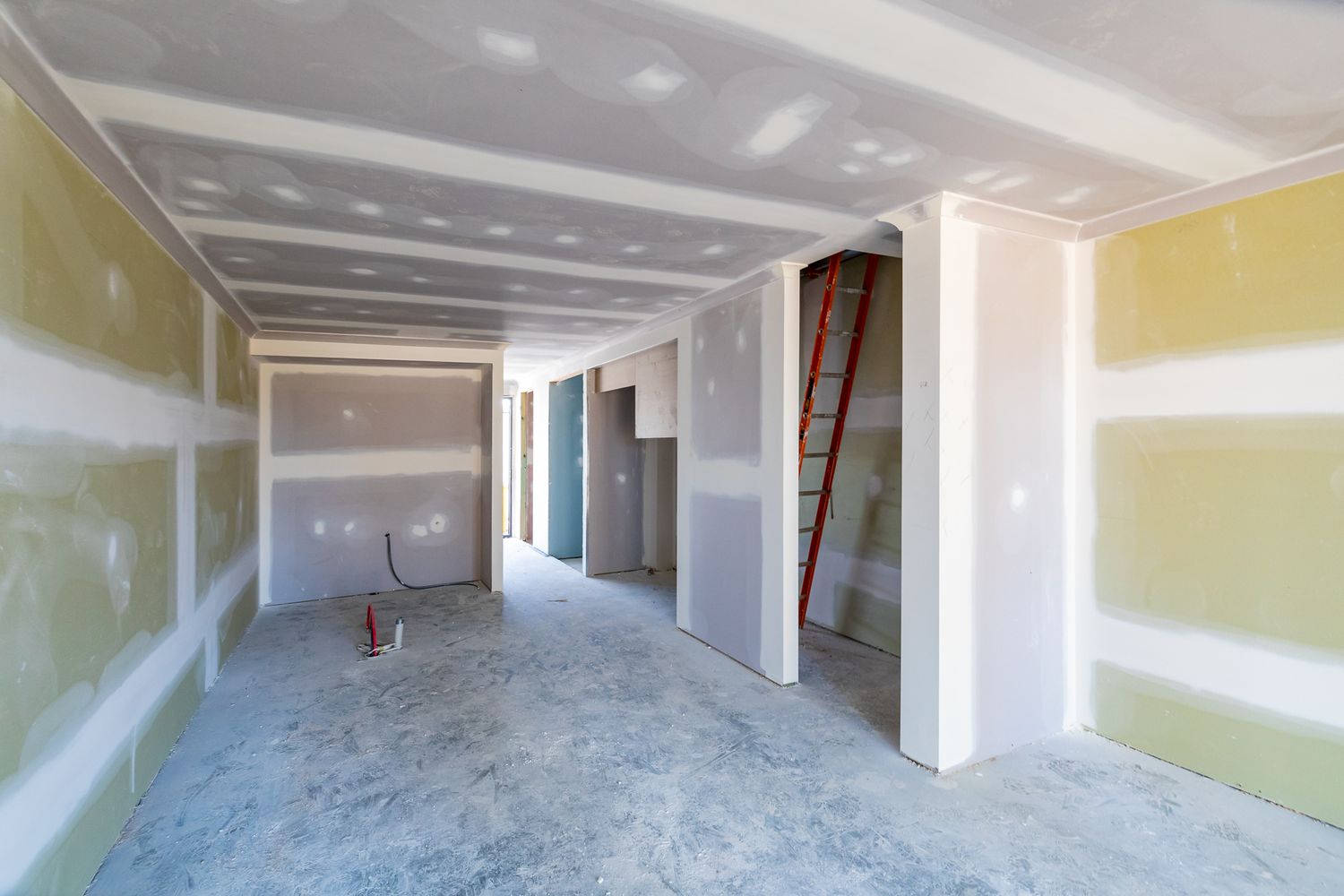 Interior of a new building under construction with plasterboard walls, exposed ceiling framing, concrete floor and a red ladder.