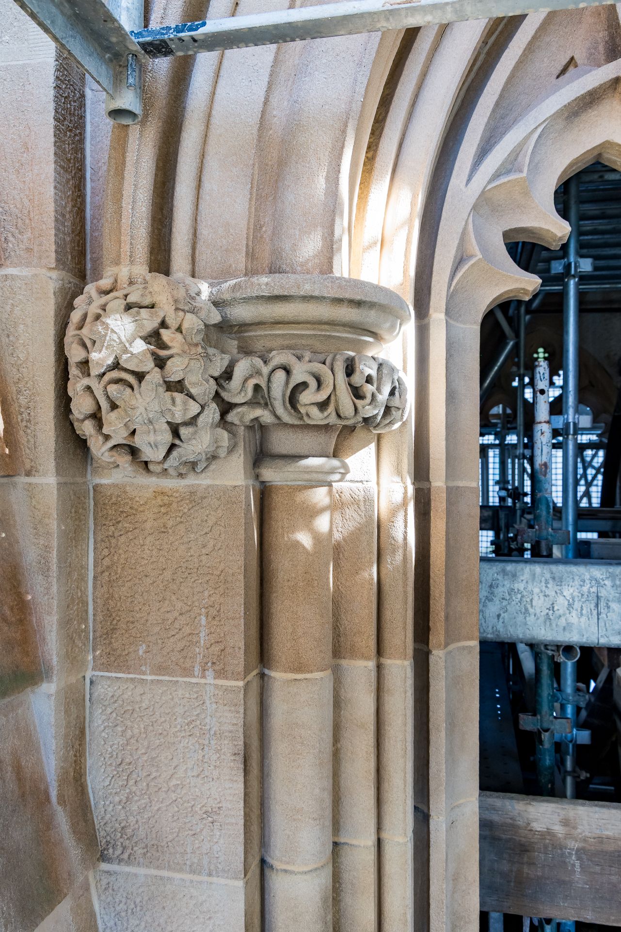 Close-up of a sandstone corbel with carved floral and scroll details beside a pointed arch and scaffolding.