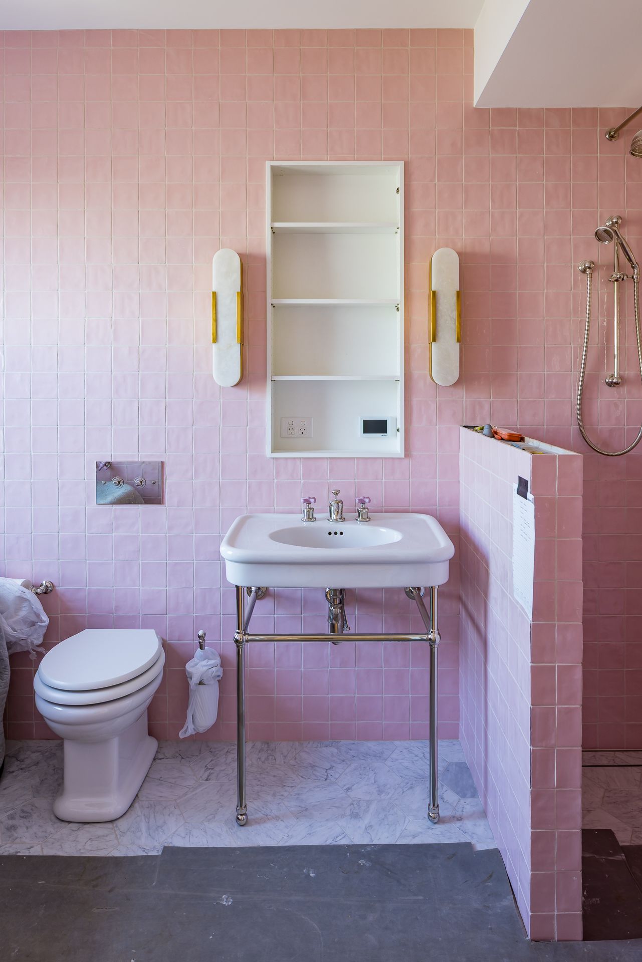 Pink tiled bathroom with a white pedestal sink, brass wall sconces, recessed medicine cabinet, vintage toilet and marble floor.