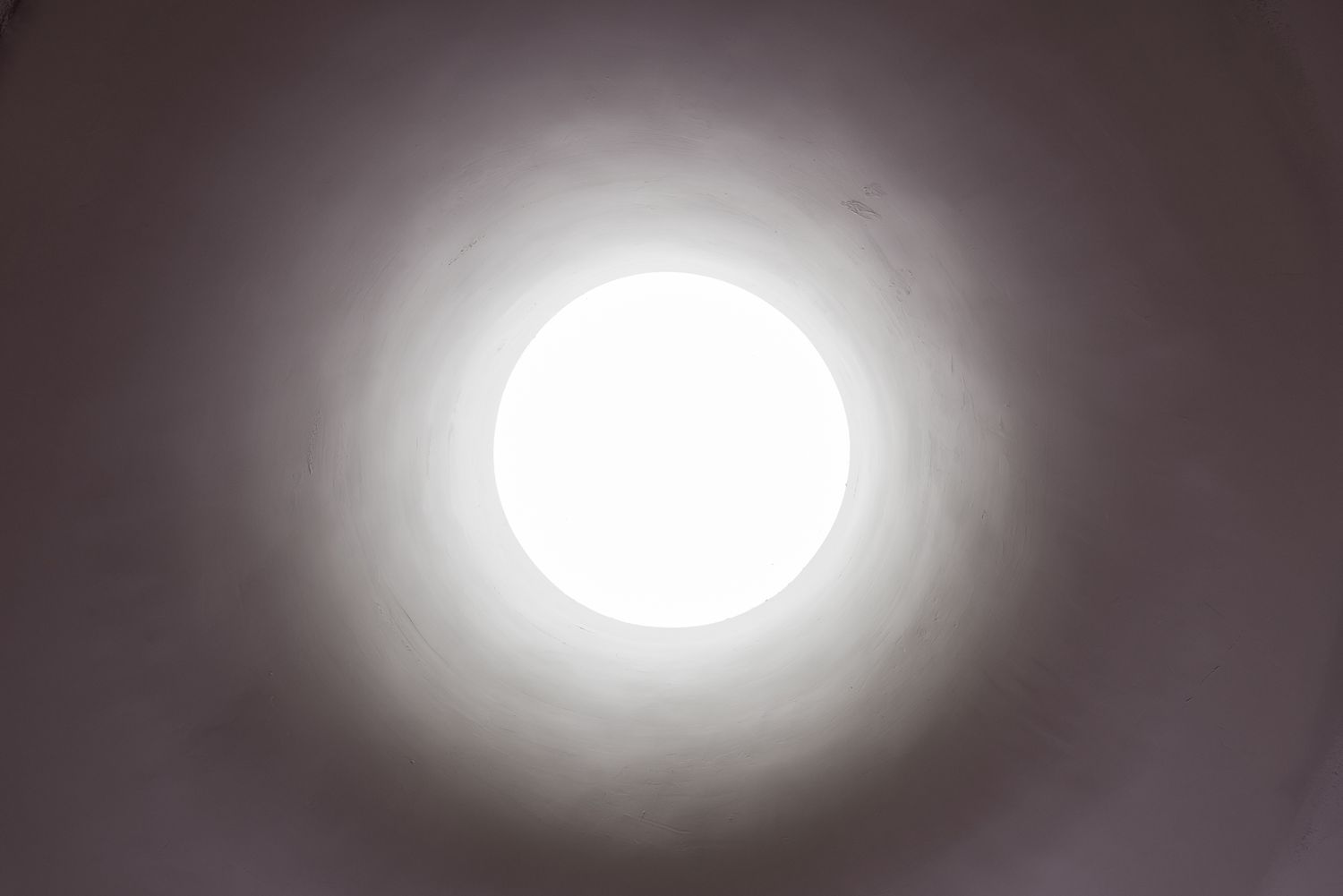 Bright circular skylight glowing in a dark ceiling, creating a soft gradient of light that fades into shadow.