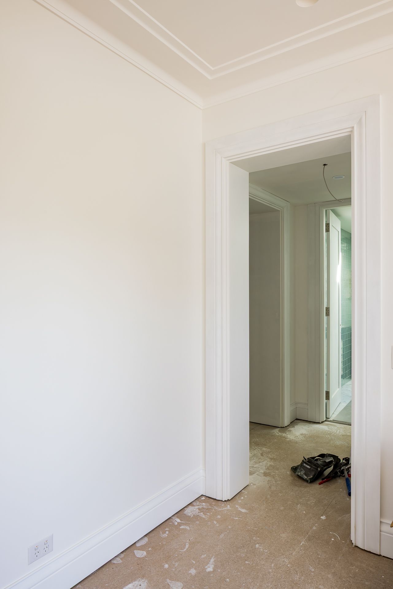 Freshly painted white room with ornate cornice and a doorway leading to a hallway with tools on the unfinished floor.