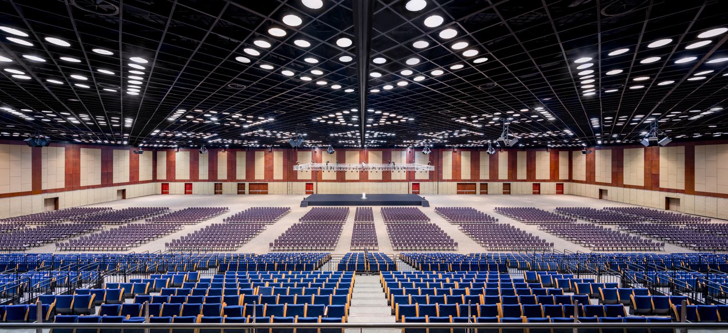 Vast convention hall interior with rows of blue chairs, a gridded ceiling of lights and a central aisle leading to the stage.