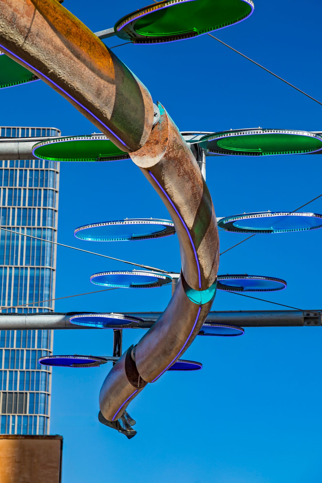 Weathered copper spiral sculpture with green and blue glowing discs on steel arms beside a glass skyscraper.