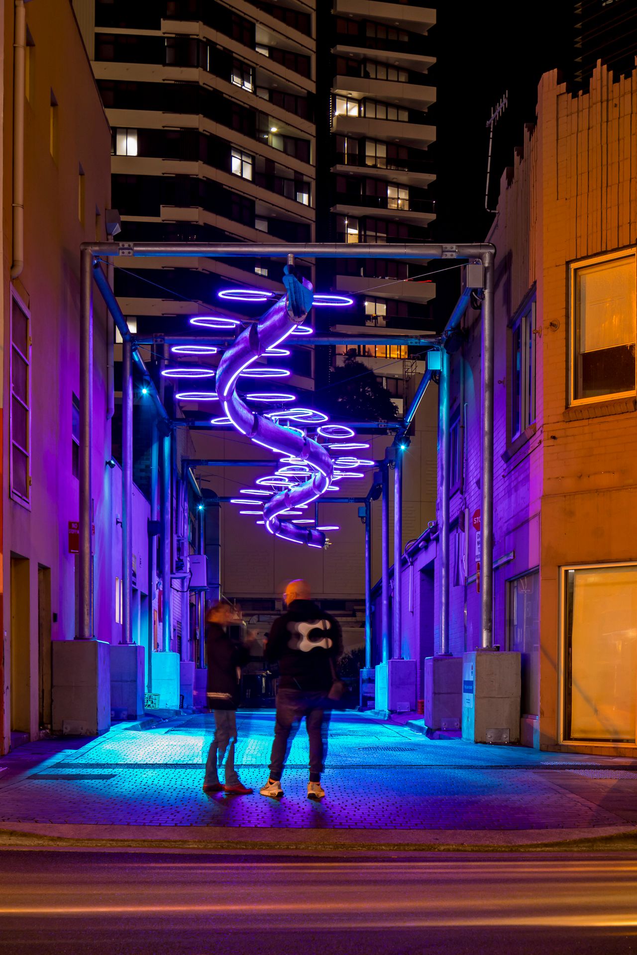 Two people stand beneath a glowing purple neon spiral sculpture in an urban laneway at night with high-rise towers behind.