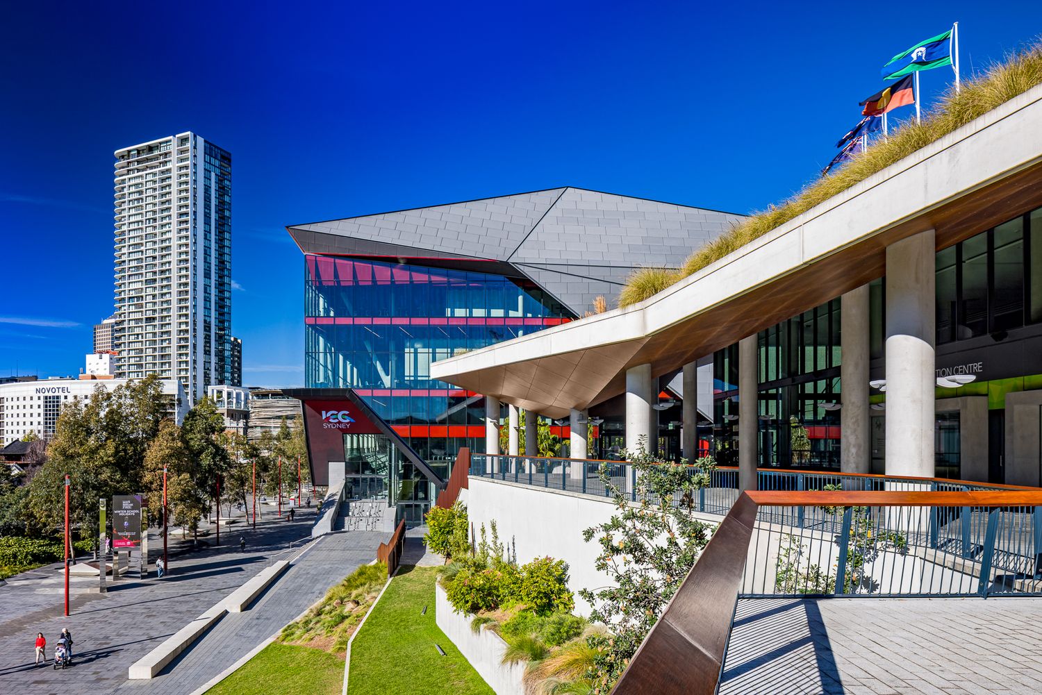 Modern convention centre with angular glass facade, tiered terraces and a high-rise tower under a clear blue sky.