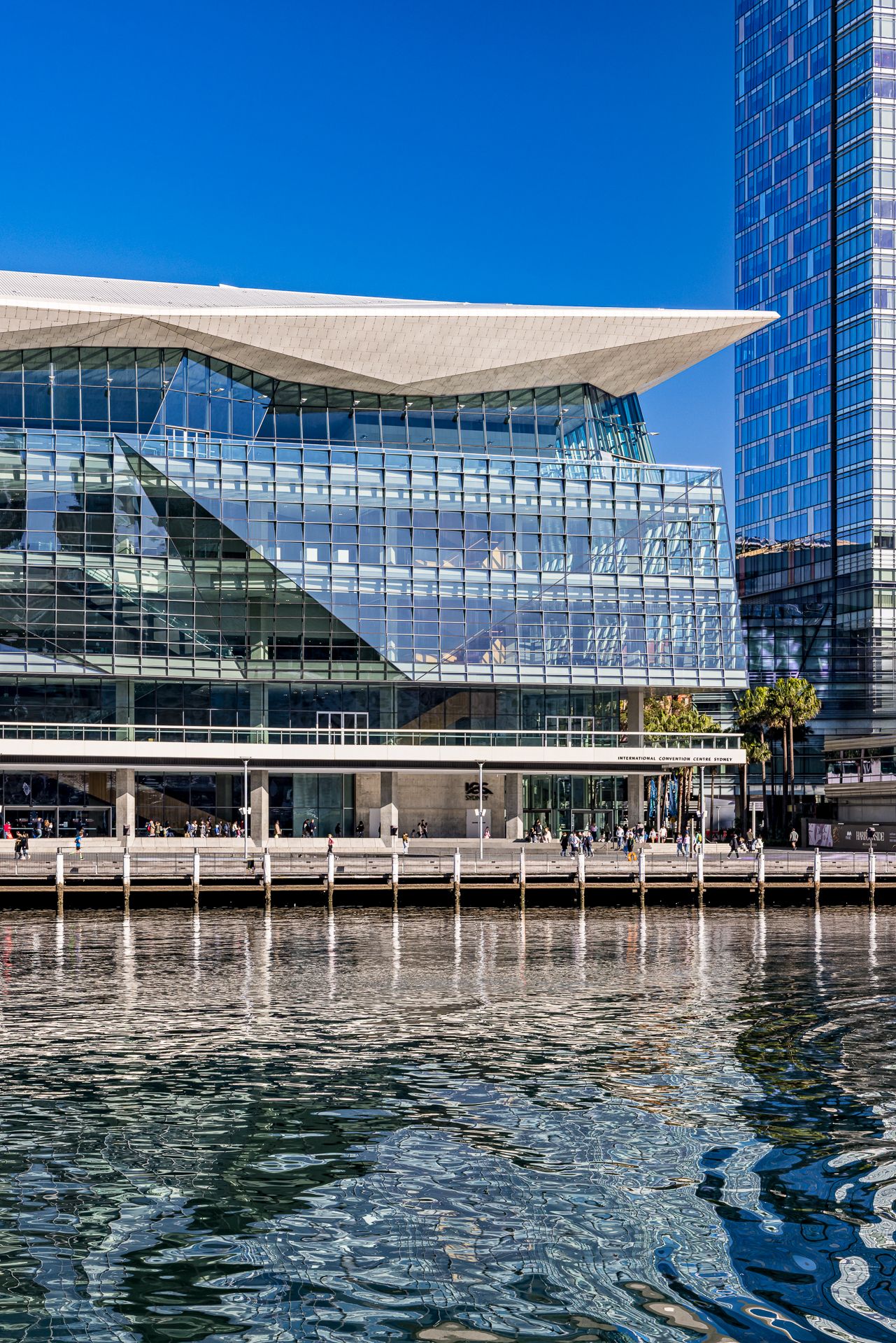Reflective glass facade of a modern convention centre with a cantilevered white roof, rippling harbour water in the foreground.
