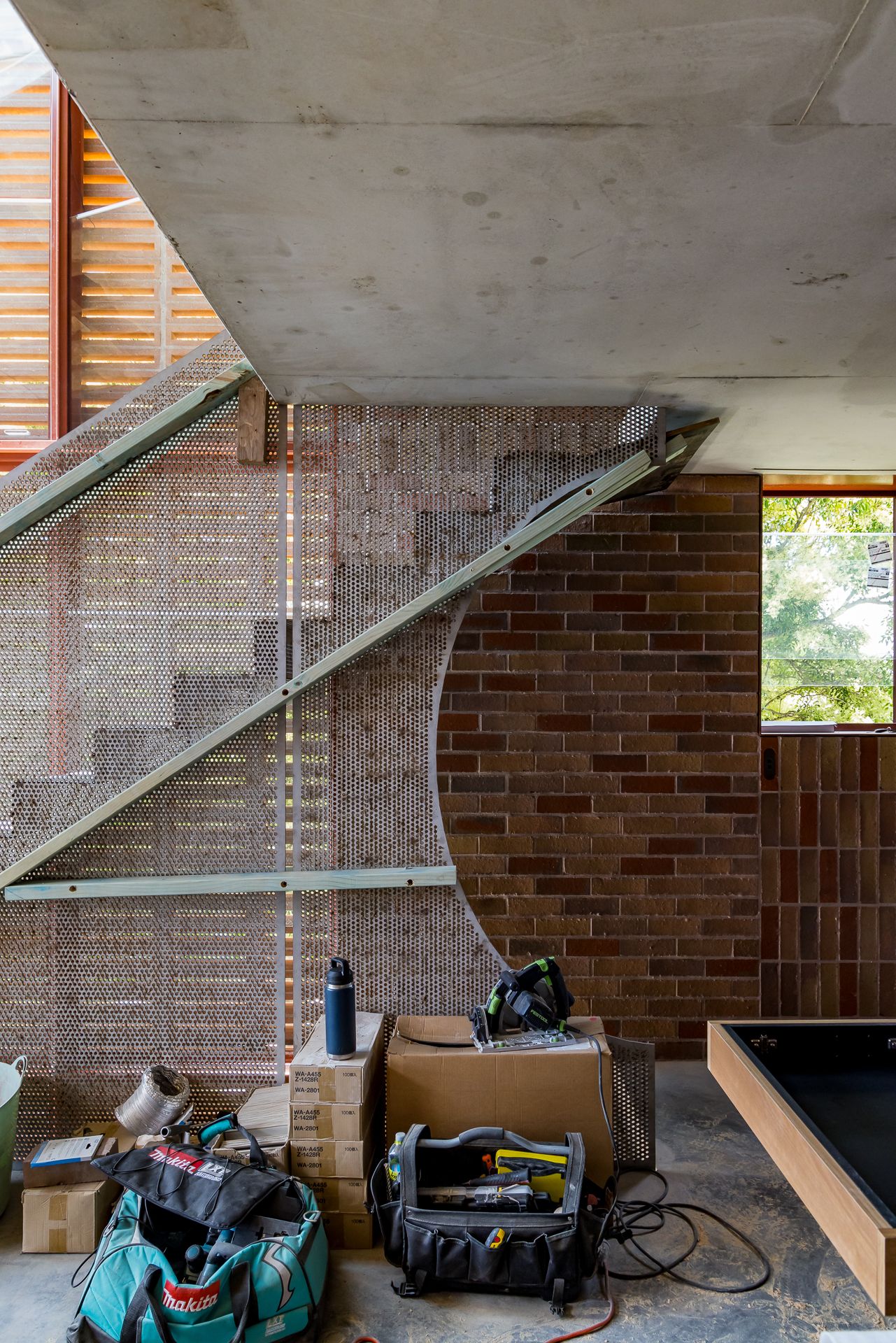 Construction site interior with tool bags and boxes below a mesh-clad stair, concrete soffit and brick wall.