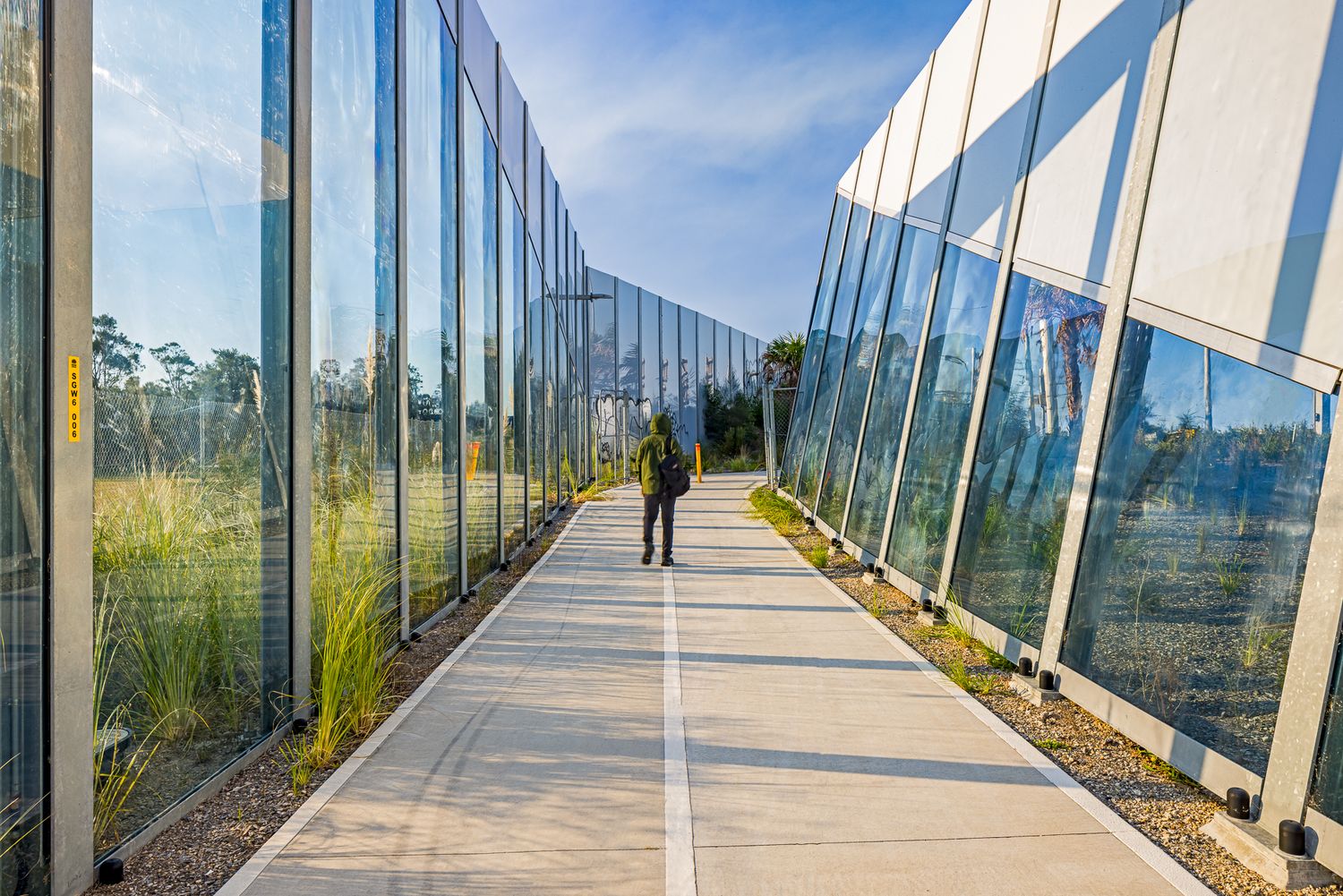 Person walking along a concrete path lined with tall glass panels and green grass beneath a bright blue sky.