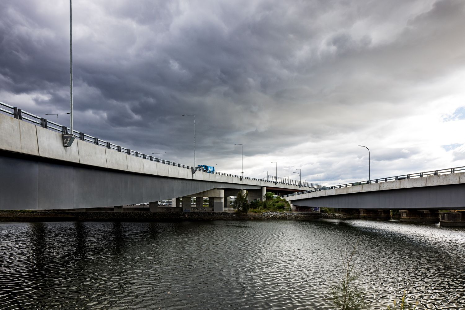 Two concrete motorway bridges spanning a calm river beneath a dramatic stormy grey sky with heavy dark clouds.