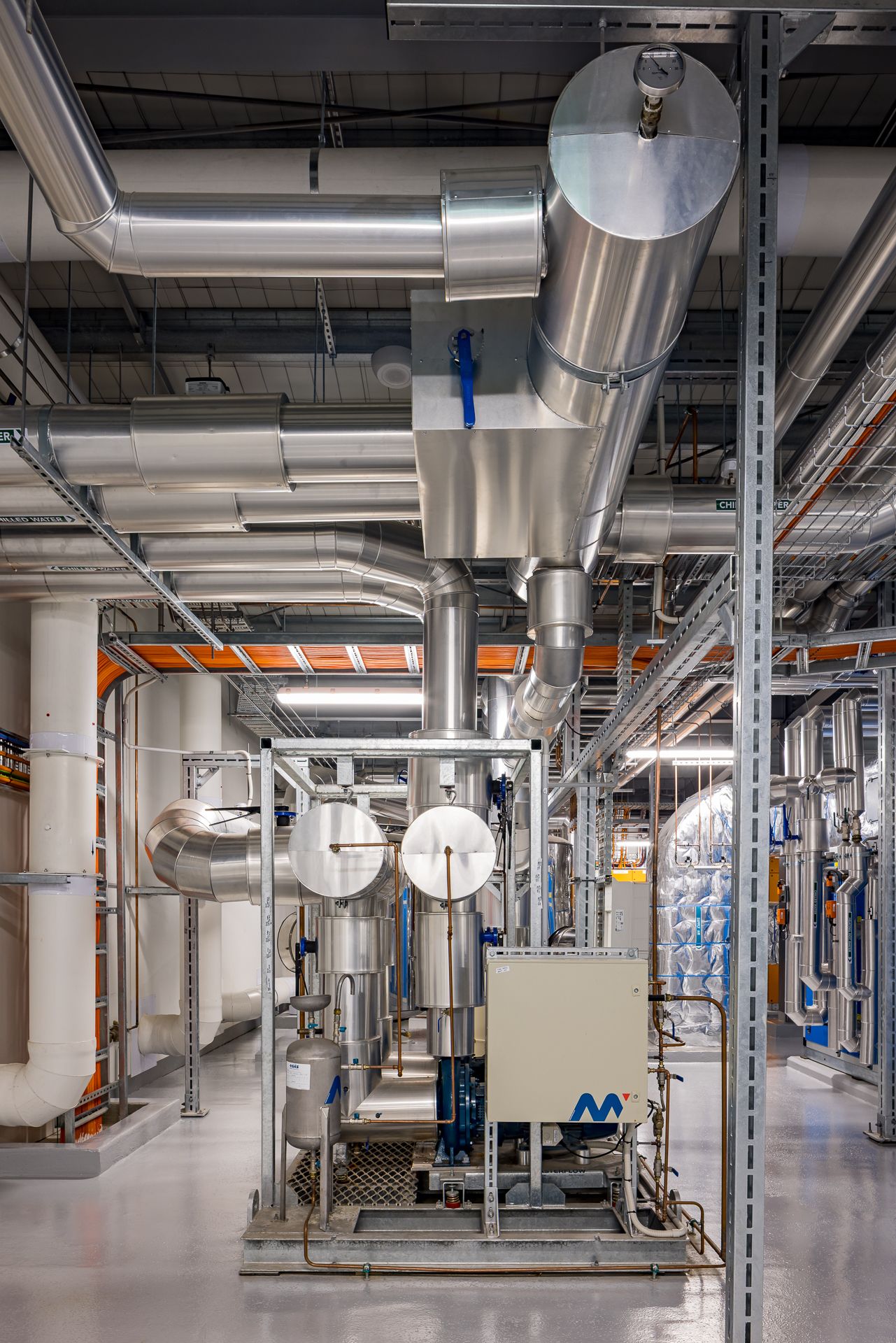 Close view of a mechanical plant room with insulated metal ducts, pumps, valves and overhead pipework supported by steel framing.
