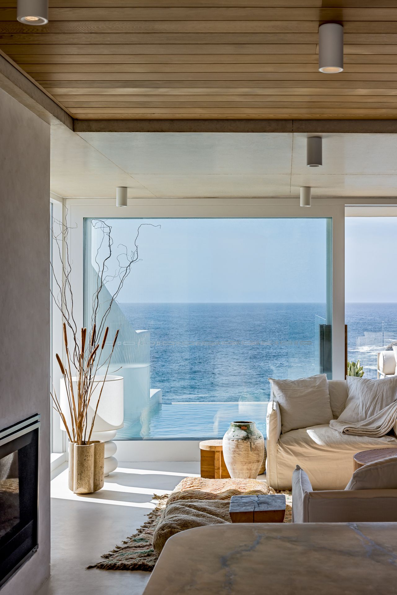 Light-filled living room with a cream sofa, wood ceiling and floor-to-ceiling glass framing a panoramic blue ocean view.