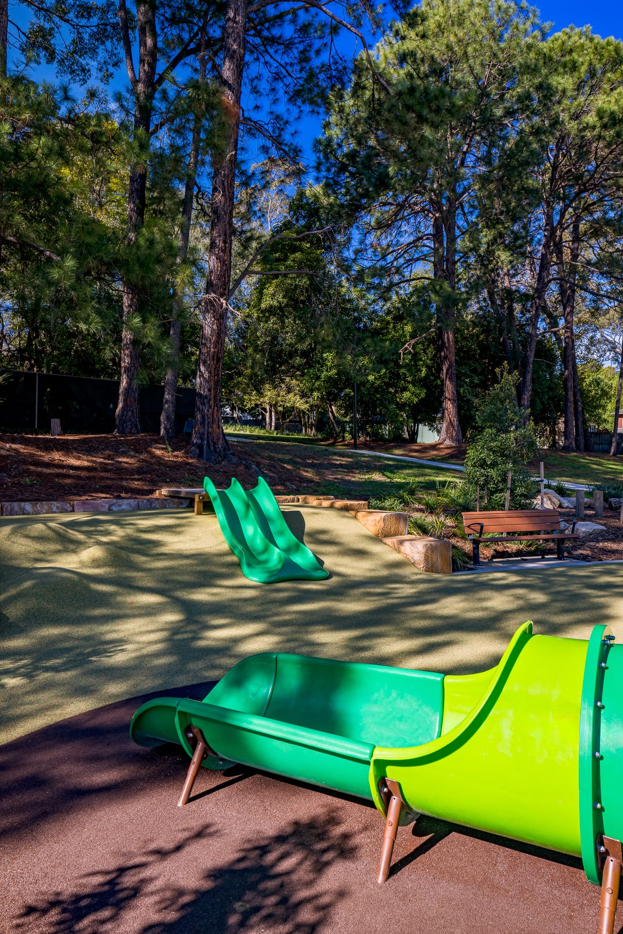 Green embankment slides on a soft rubber playground surface beside a wooden bench, set among tall pine trees in a shady park.