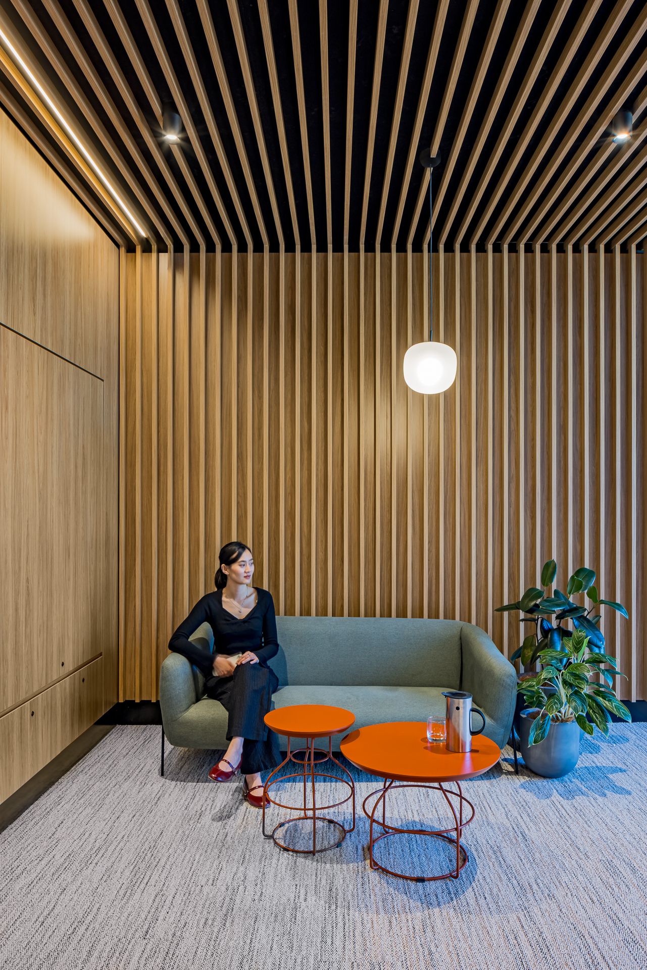 Woman seated on a green sofa in a modern waiting area with vertical timber slat walls, orange side tables and a pendant light.