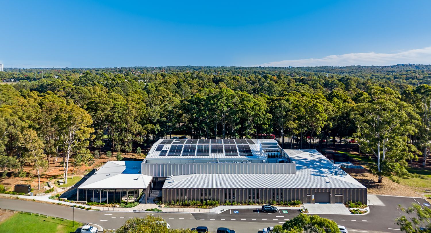 Aerial view of a modern building with rooftop solar panels surrounded by dense bushland, a car park and an access road.