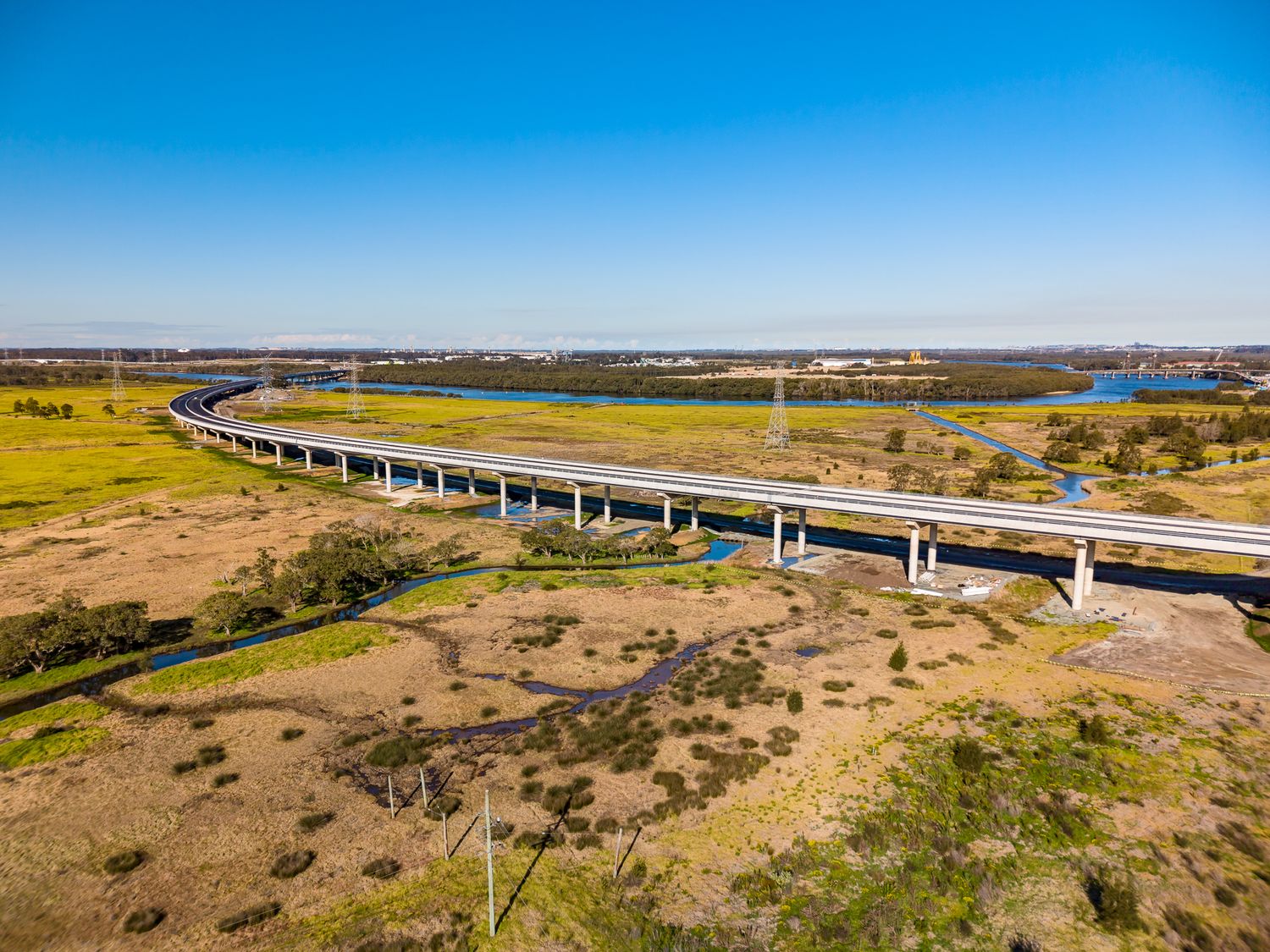Aerial view of a long elevated highway bridge crossing wetlands and a shallow creek beneath a clear blue sky.