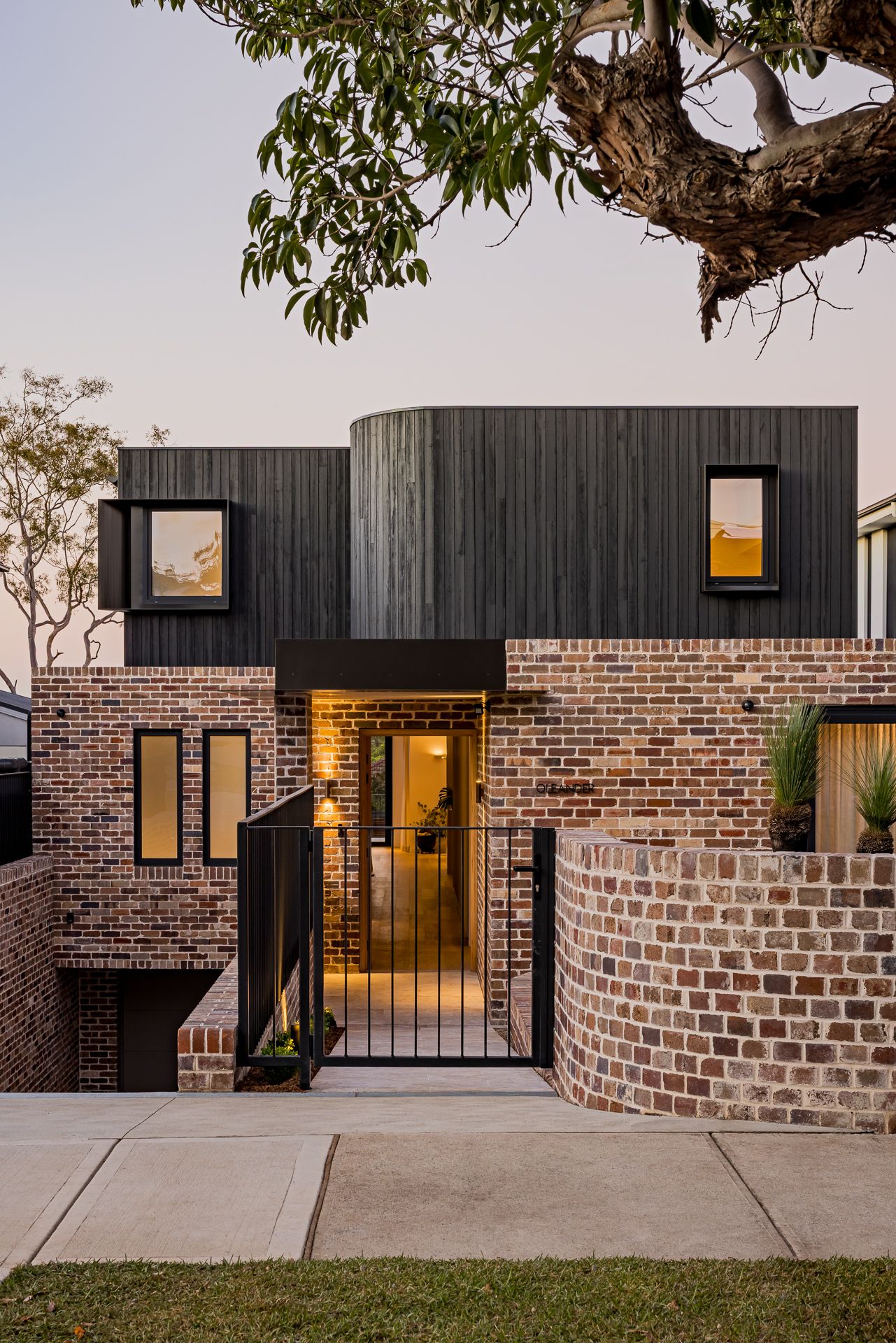 Curved black timber upper storey above a rustic brick facade with a curved front wall, glowing entry and steel gate at twilight.