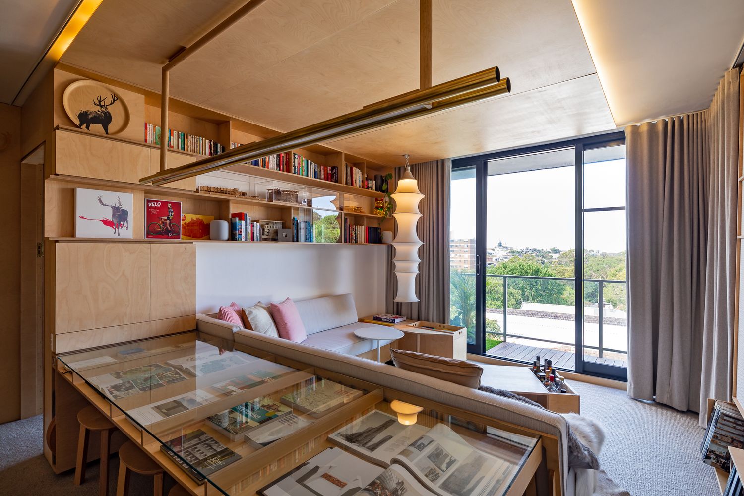 Open-plan plywood apartment with a glass display table, built-in bookshelves, pendant lamp, and balcony views.