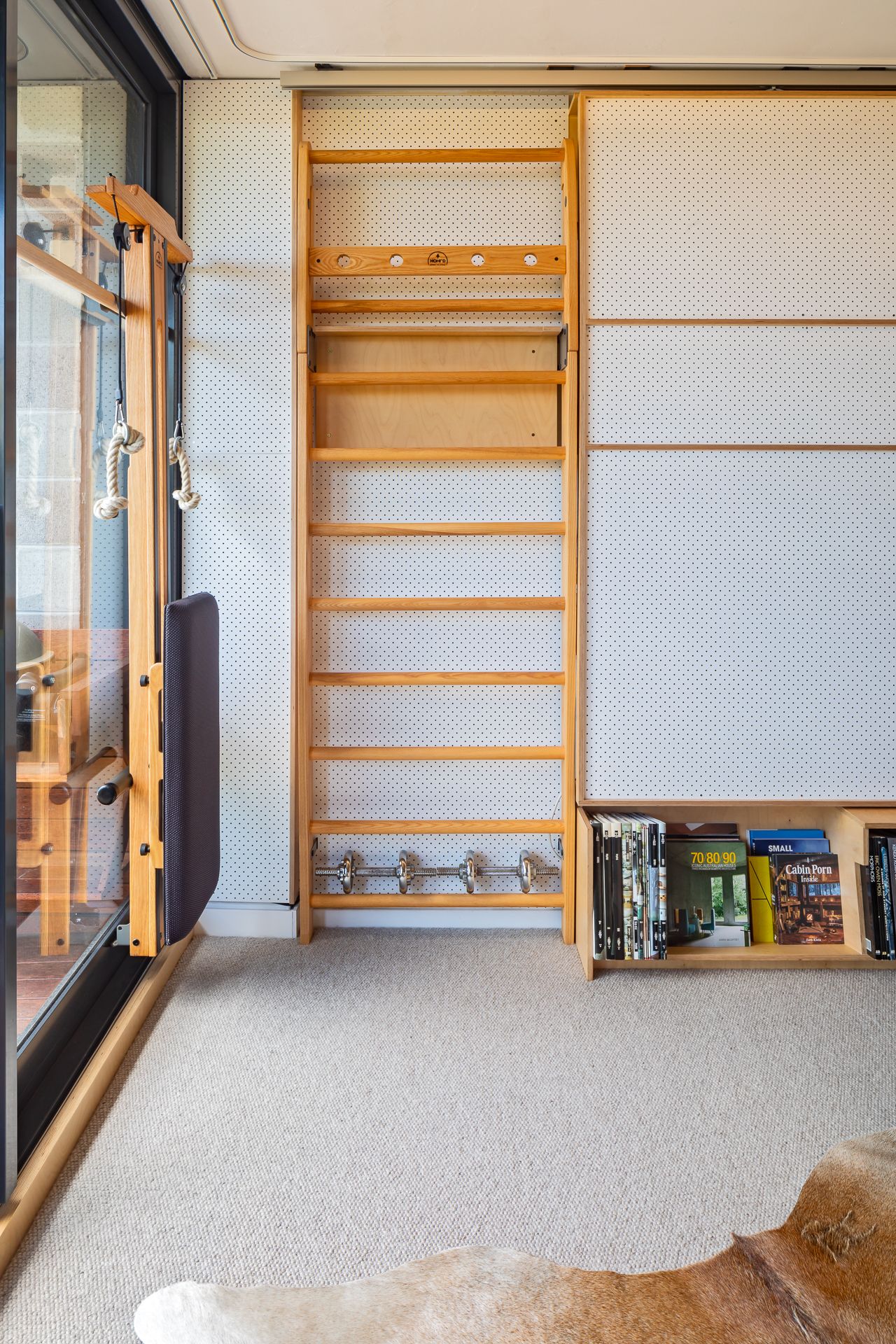 Wooden swedish ladder wall bars with hooks and a folded padded bench in a home gym with beige carpet and cowhide rug.