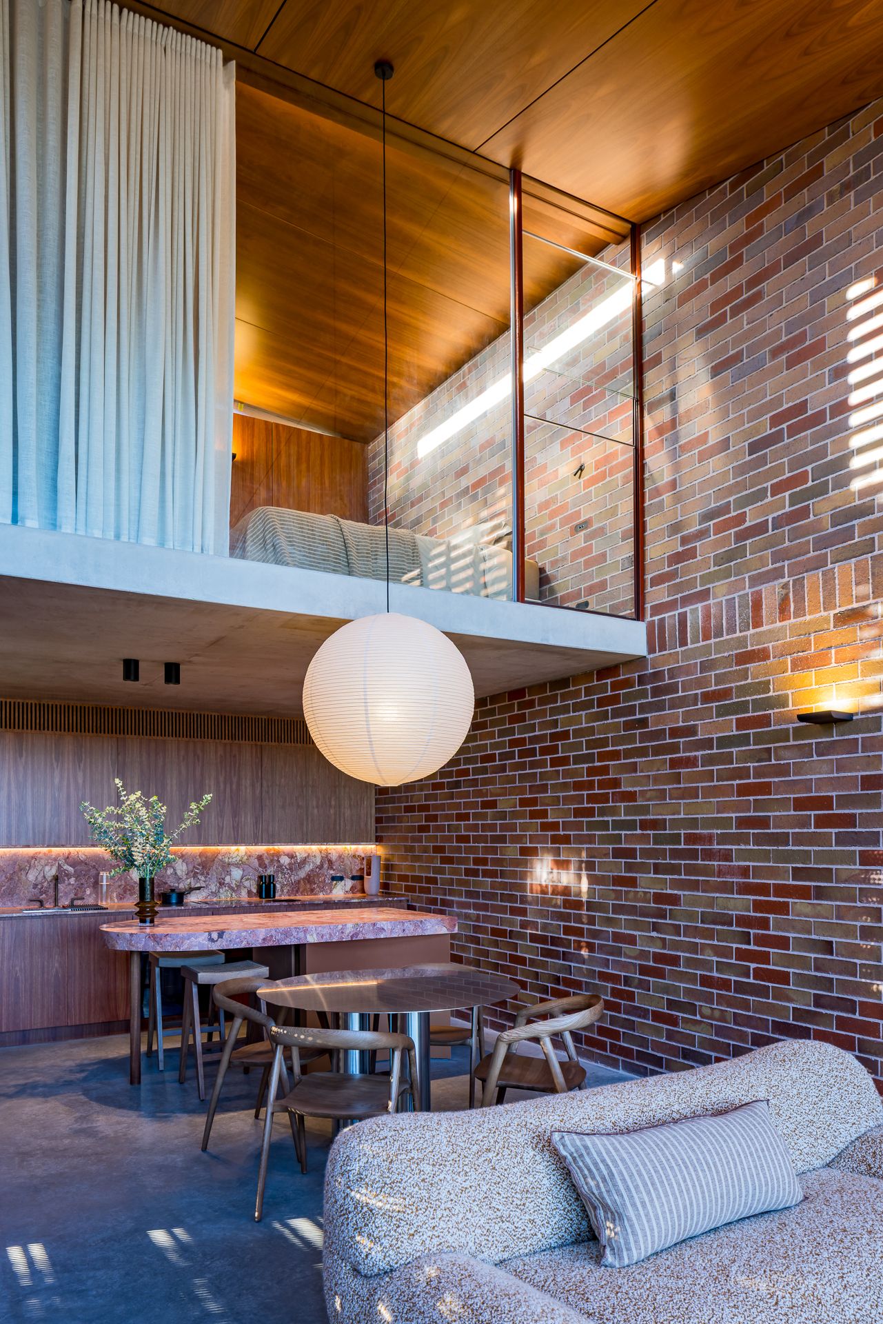 Double-height interior with exposed brick, timber ceilings, a paper lantern, a marble kitchen bench and a mezzanine bedroom.