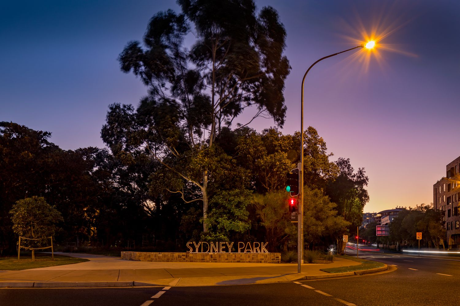 Sydney Park entrance sign and tall trees at twilight, with a glowing street lamp and traffic lights along the quiet road.