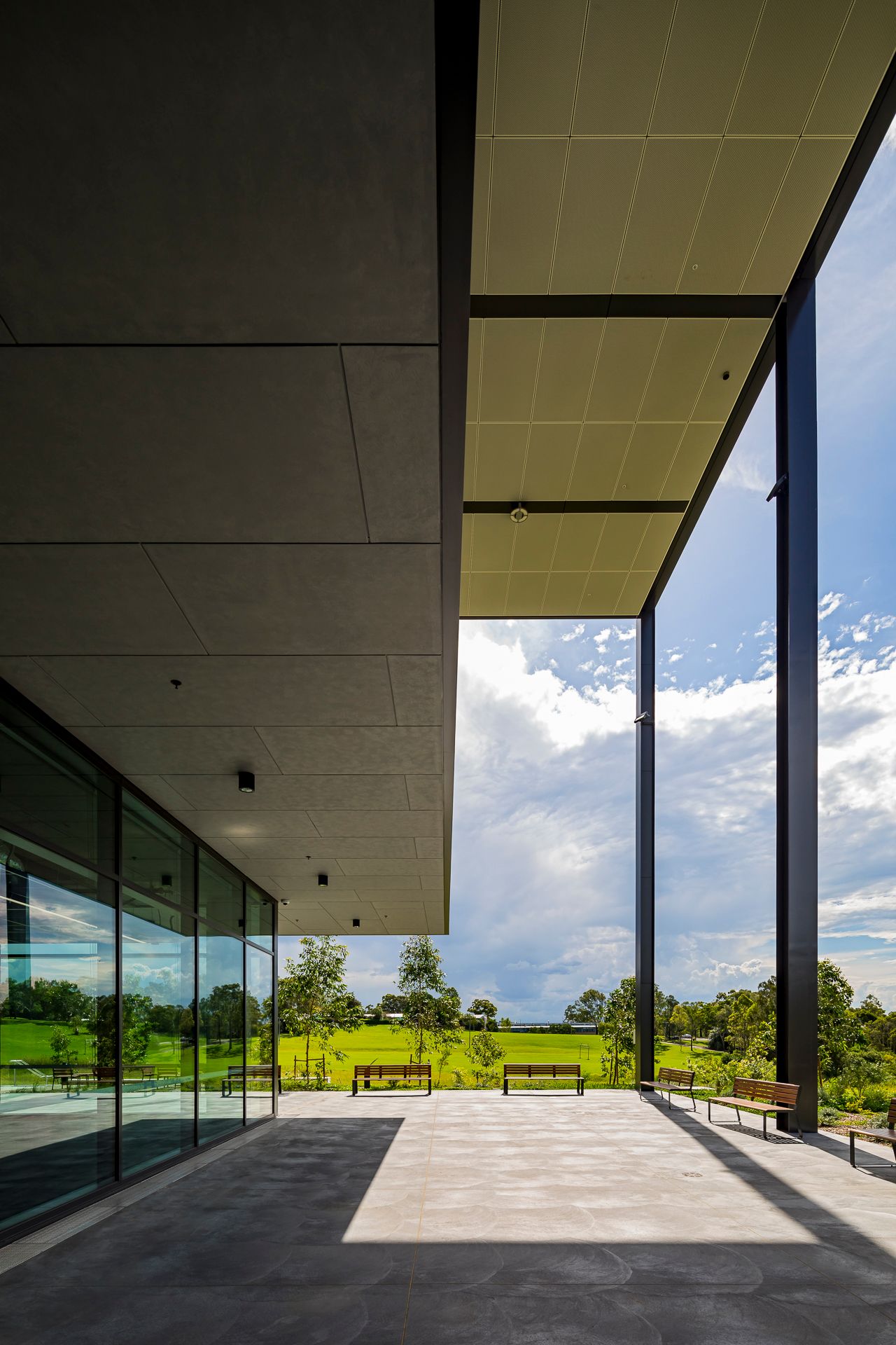 Covered outdoor terrace with steel columns, glass walls and park benches overlooking a green lawn and blue sky.