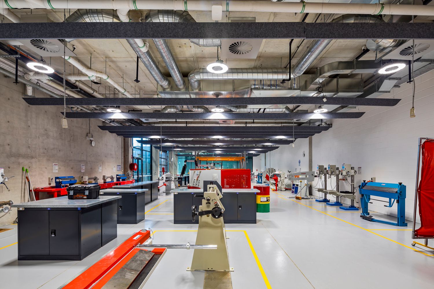 Spacious industrial workshop with exposed ceiling ductwork, recessed lighting, workbenches, and machine tools on a polished concrete floor.