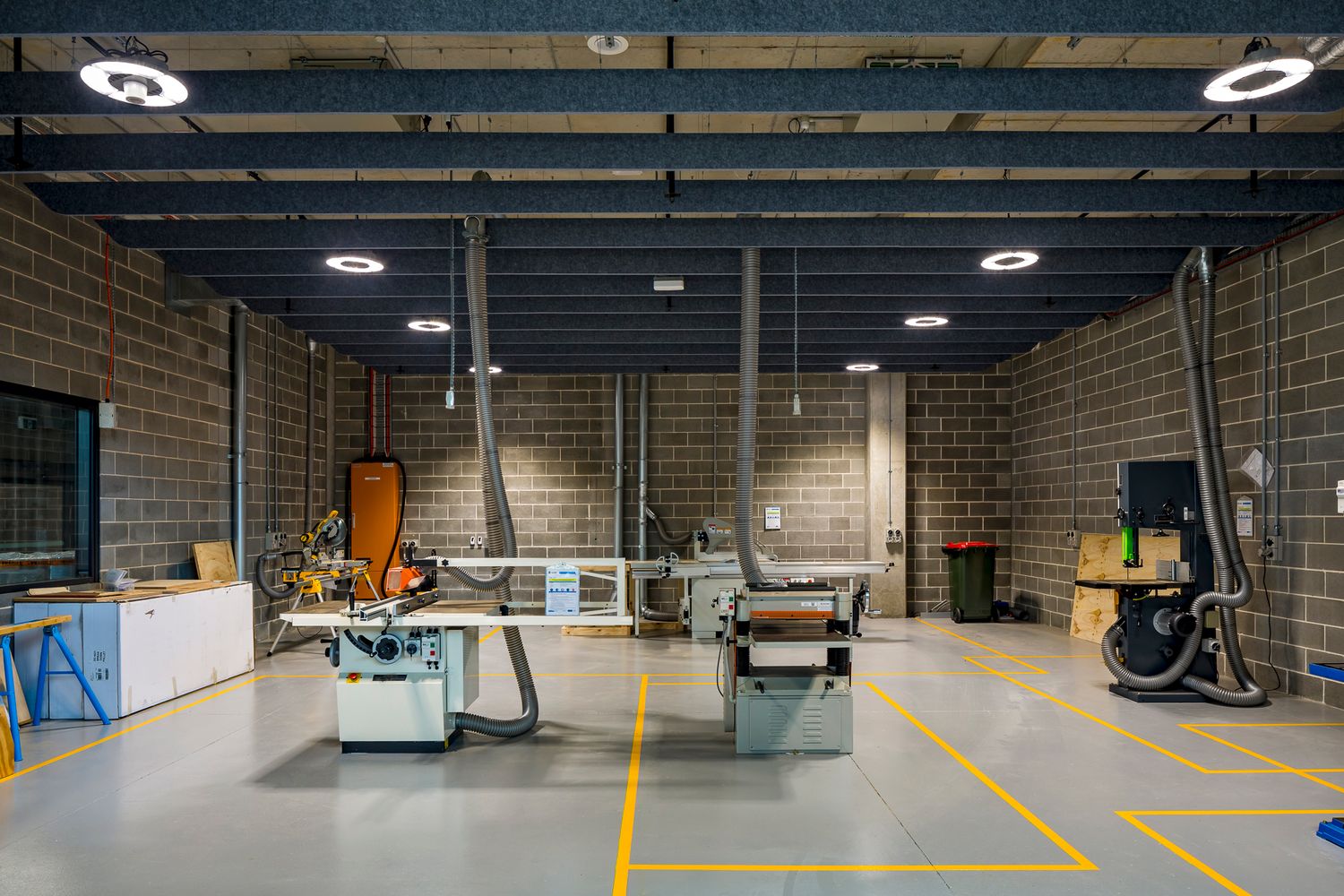 Spacious industrial workshop with brick walls, yellow floor markings and woodworking machinery under a dark exposed ceiling.