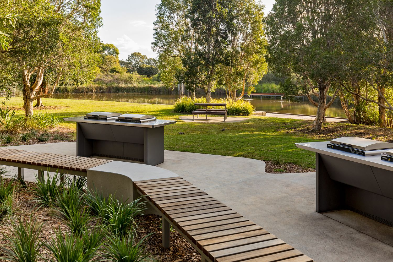 Timber boardwalk and concrete barbecue stations in a landscaped park with lawn, native trees and waterside views.