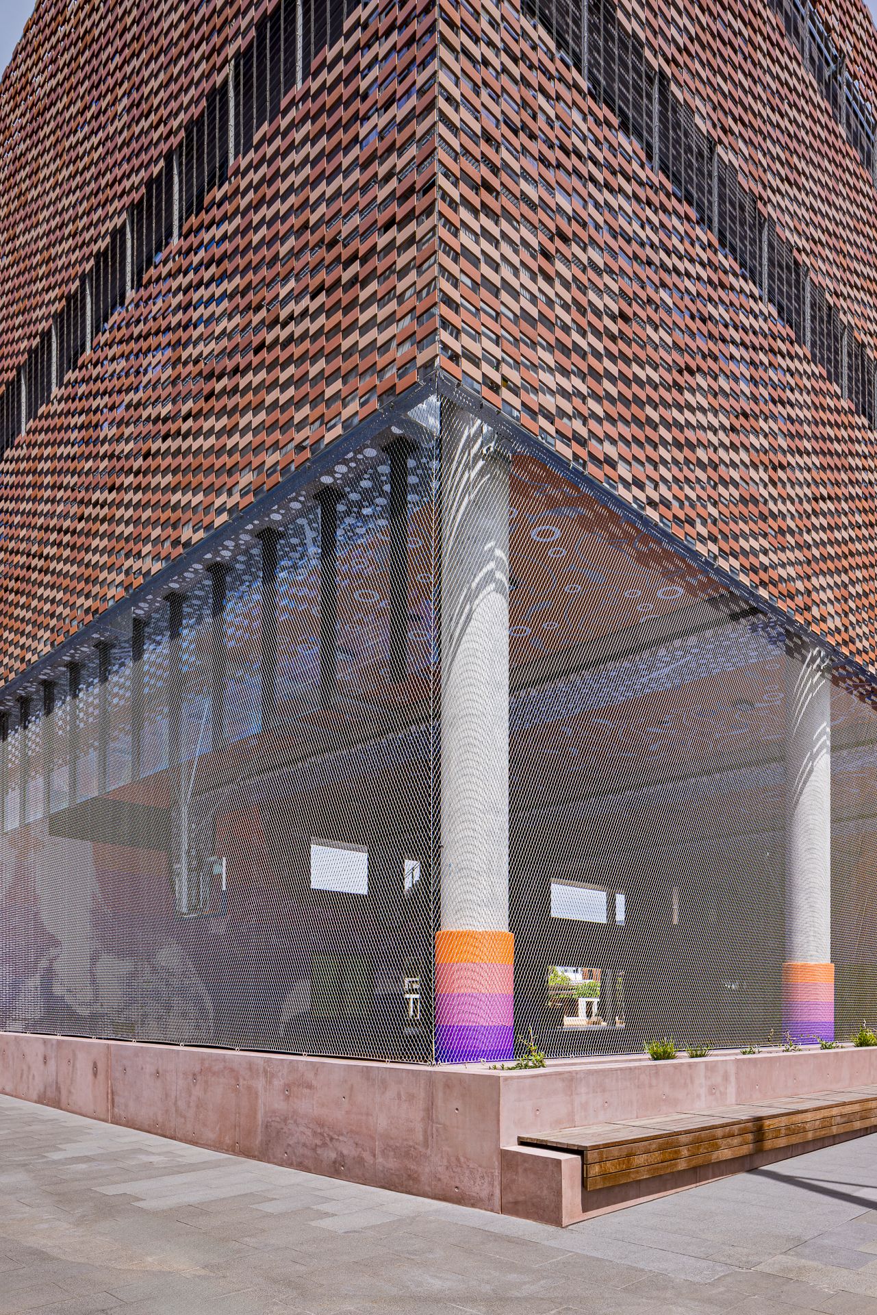 Modern building corner with textured terracotta brick facade, translucent mesh screen and colorful striped columns above a wooden bench.