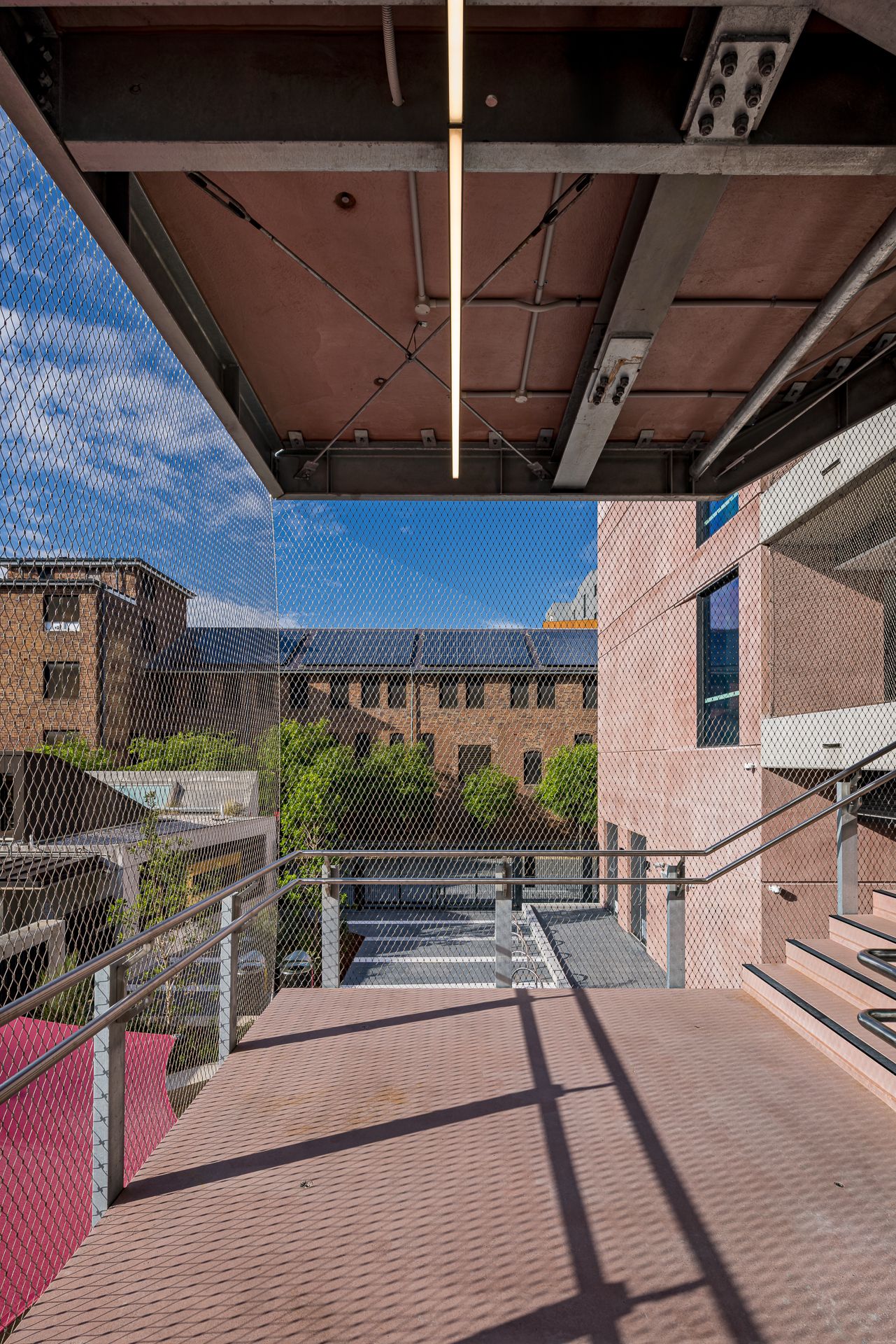 Open-air walkway with red-toned flooring and steel beams overhead, mesh screening, and brick buildings with solar panels beyond.