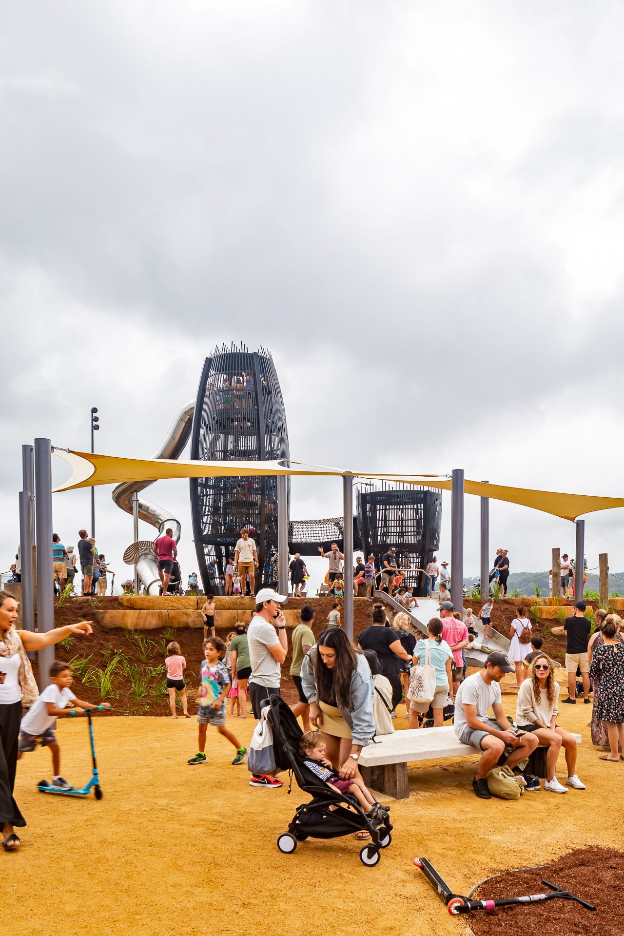 Children, parents and strollers fill a sandy playground shaded by yellow sails, with tall black climbing towers and a slide behind.
