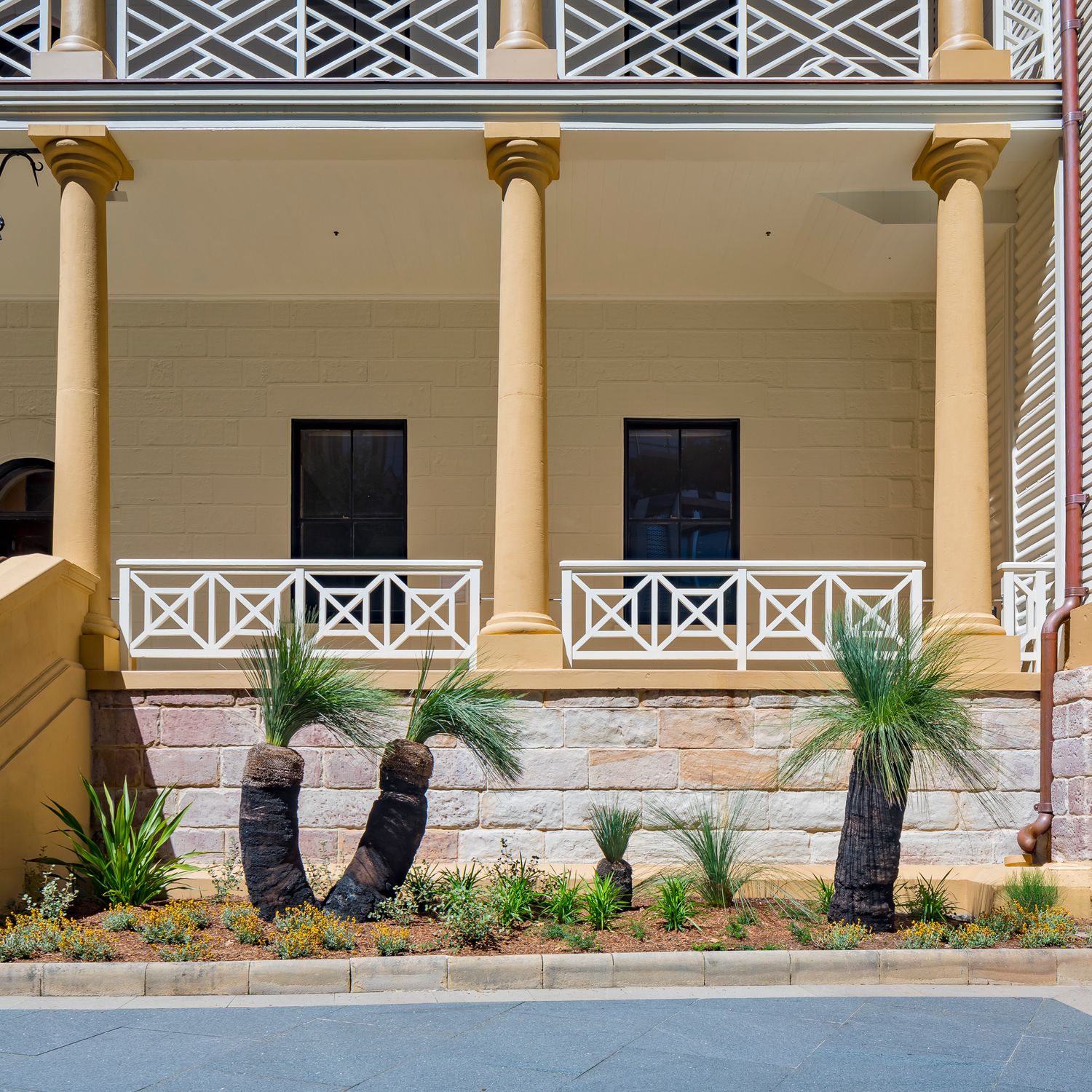 Historic sandstone building with a columned veranda, white lattice railings, and grass trees planted in a landscaped garden bed.