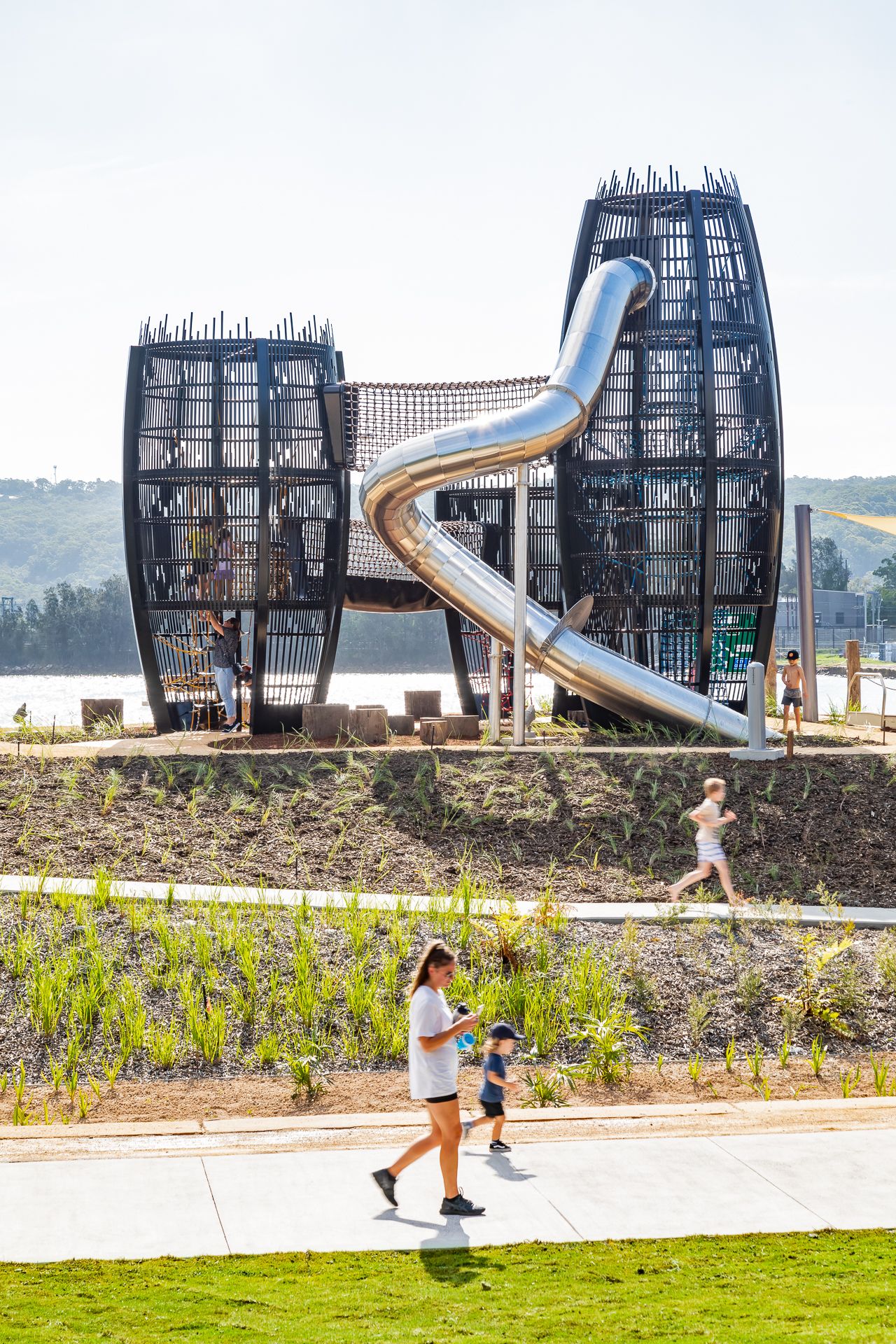 Two giant black woven lantern-shaped play towers connected by a silver spiral slide overlook a waterfront park as families walk past.