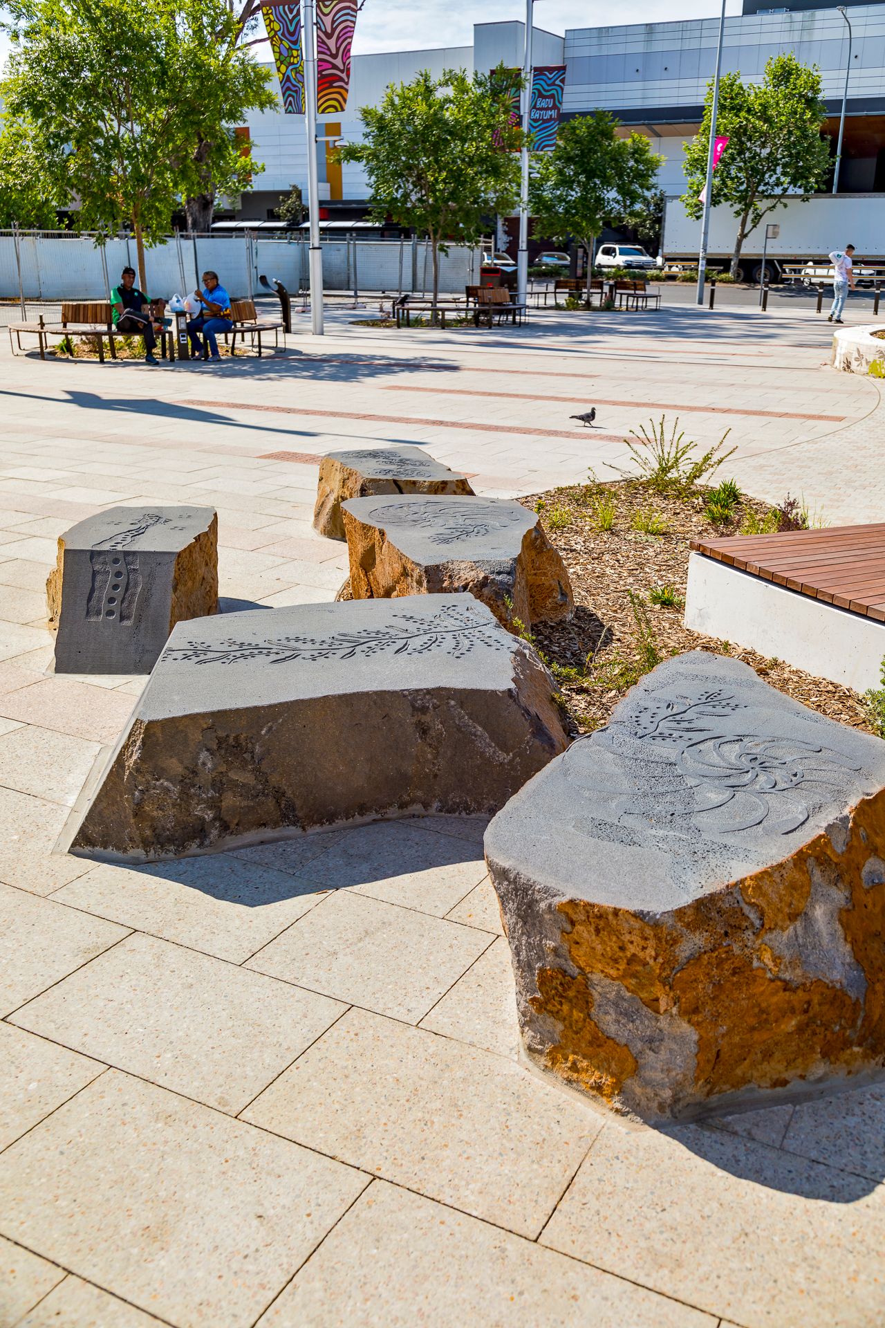 Carved sandstone boulders with etched patterns serve as public seating in a paved plaza with garden beds, trees and distant shops.