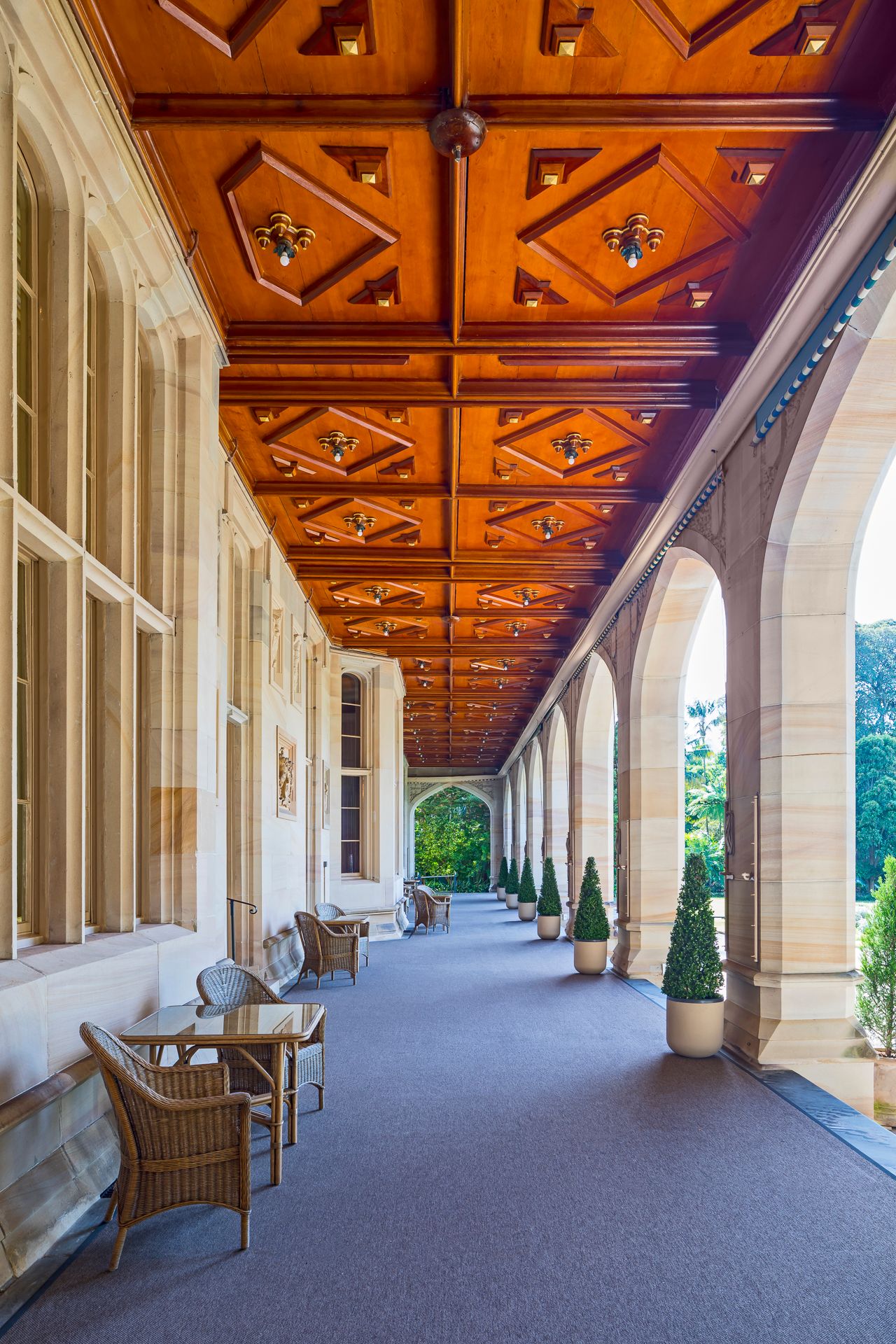 Covered stone arcade with ornate coffered timber ceiling, wicker chairs, and potted topiary trees overlooking a sunny garden.