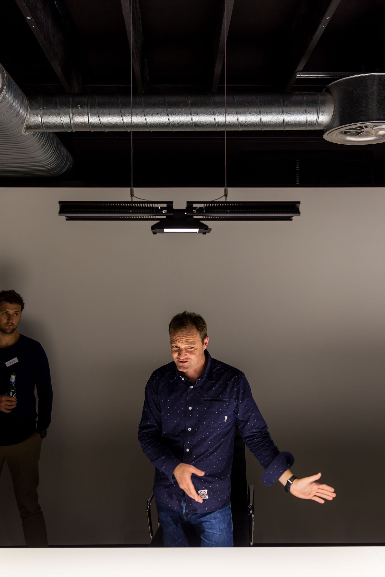 A man in a navy shirt gestures while speaking in a dimly lit studio space with another man standing behind him.