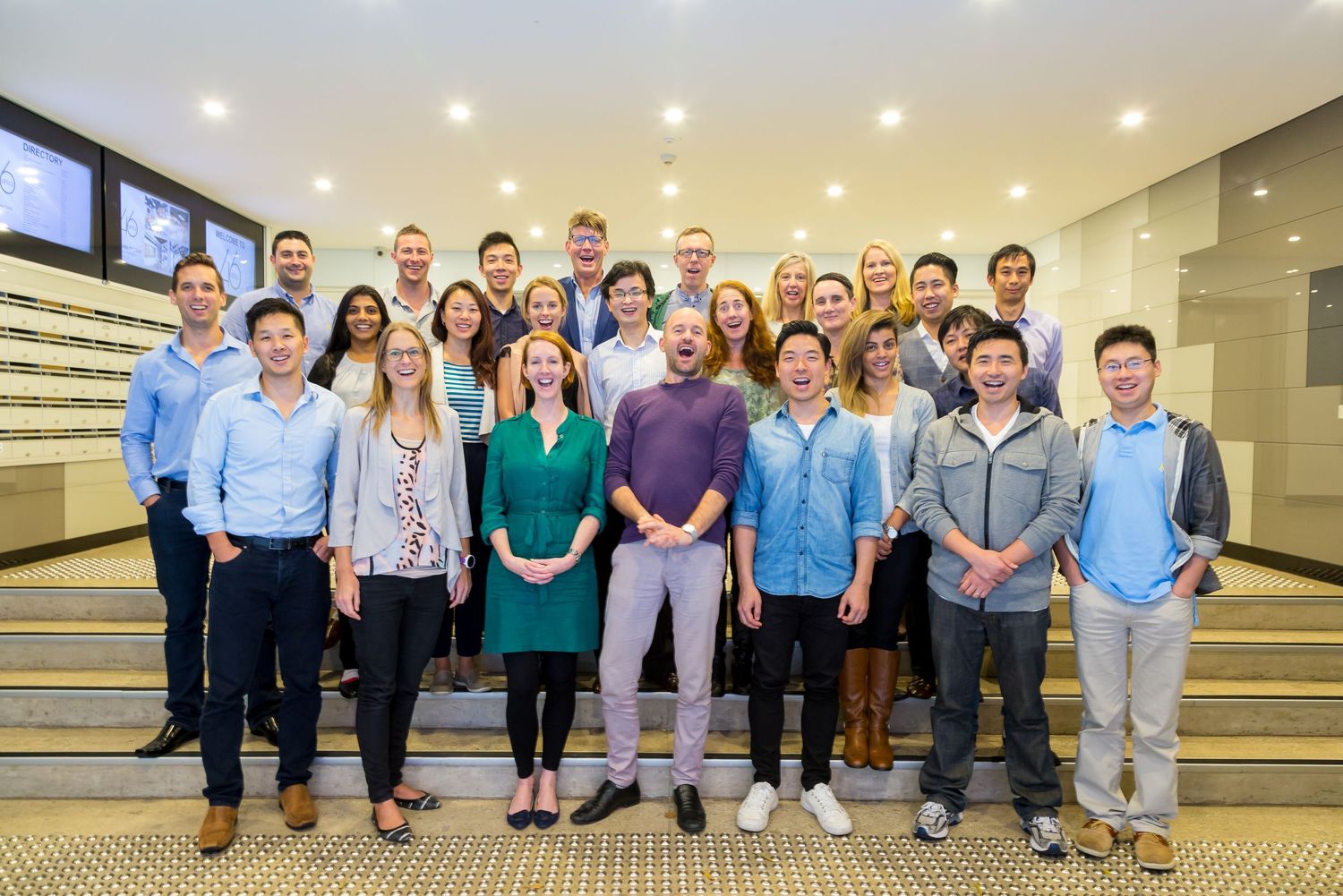 A large group of smiling colleagues in smart casual clothing pose together on wide steps in a modern office lobby.