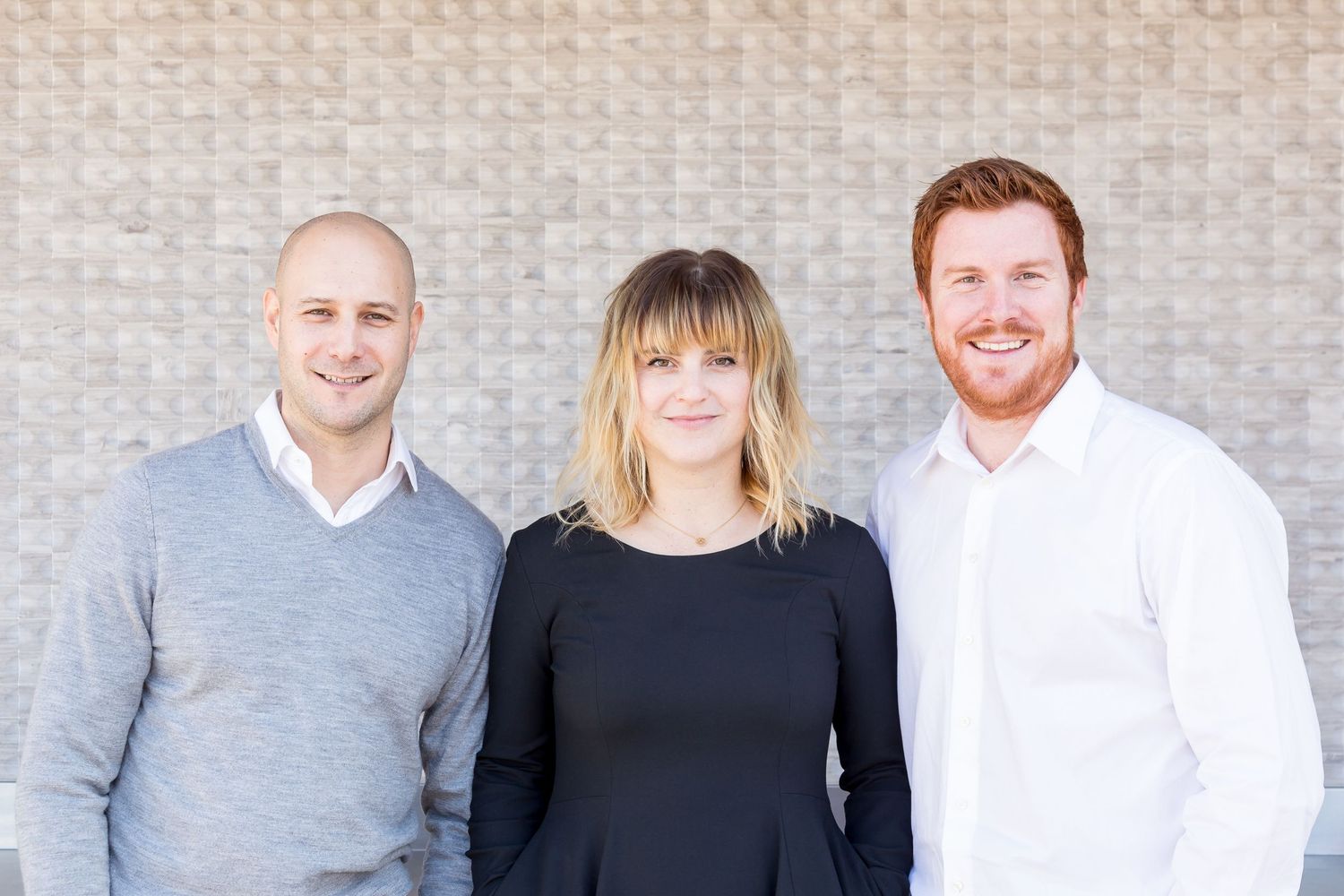 Two men and a woman with blonde hair pose smiling together in front of a light brick wall in a friendly team portrait.