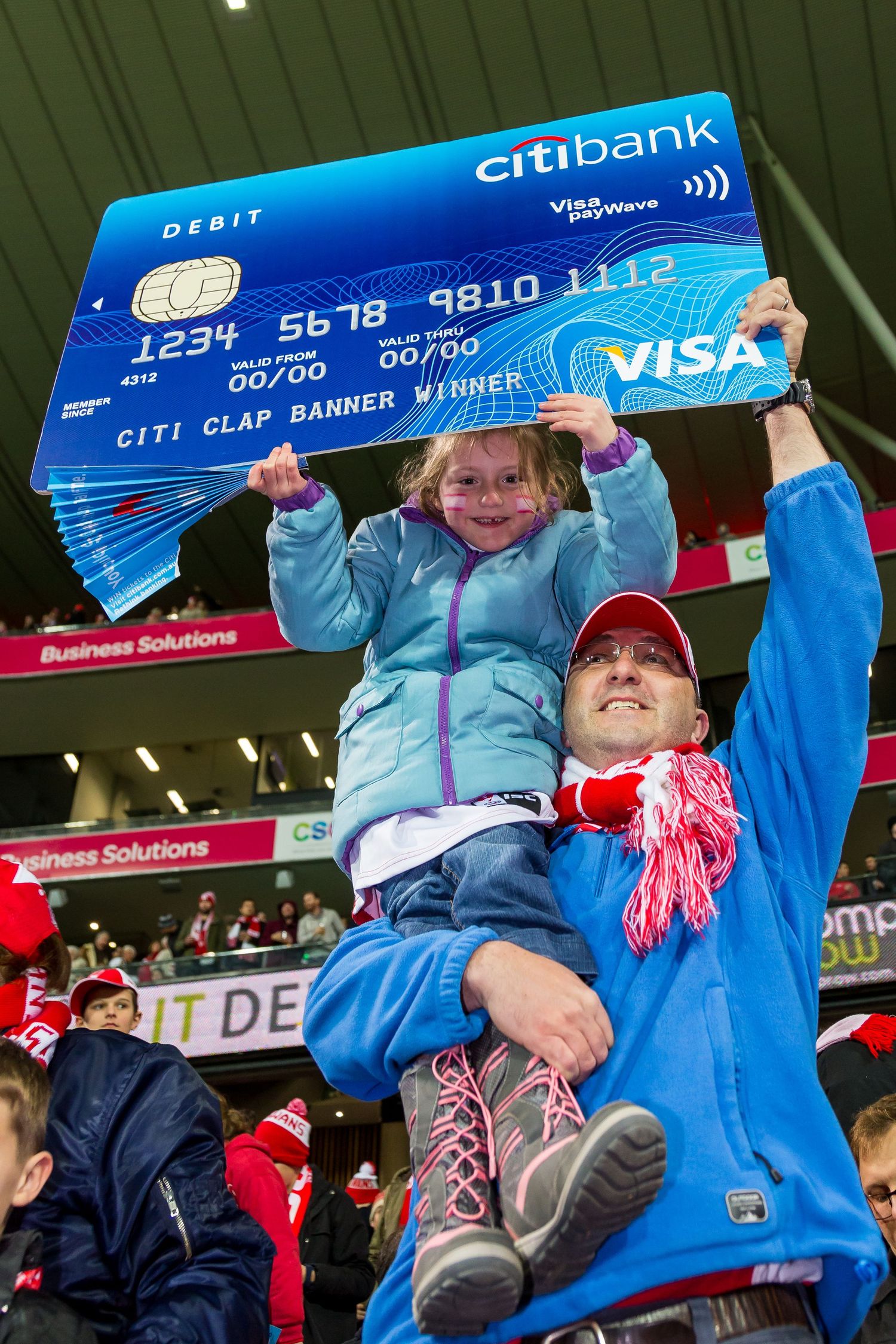 A smiling man in a red cap lifts a little girl who holds an oversized Citibank Visa debit card banner at a crowded stadium.