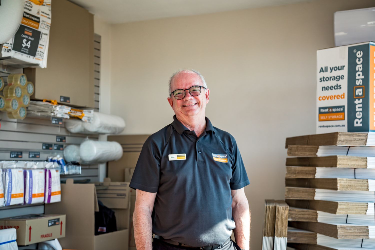 Smiling man in glasses and a dark polo shirt stands in a Rent a Space storage facility beside stacked flattened boxes and packing supplies.